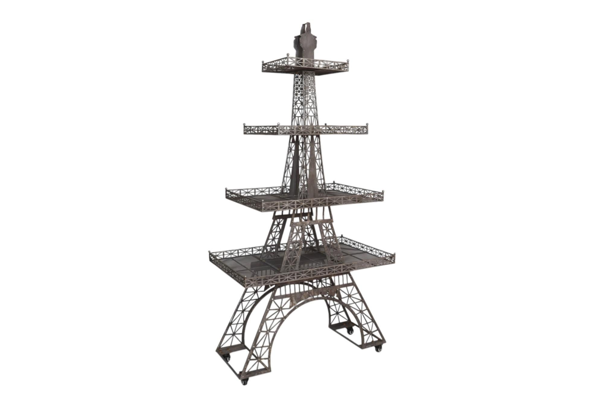 A sculptural etagere, shaped to give the look of the Eiffel Tower. The metal lattice frame supports four rectangular shelves, each with a fence like gallery rail, and ball finial topped corners. Moves easily on wheel castors with locking