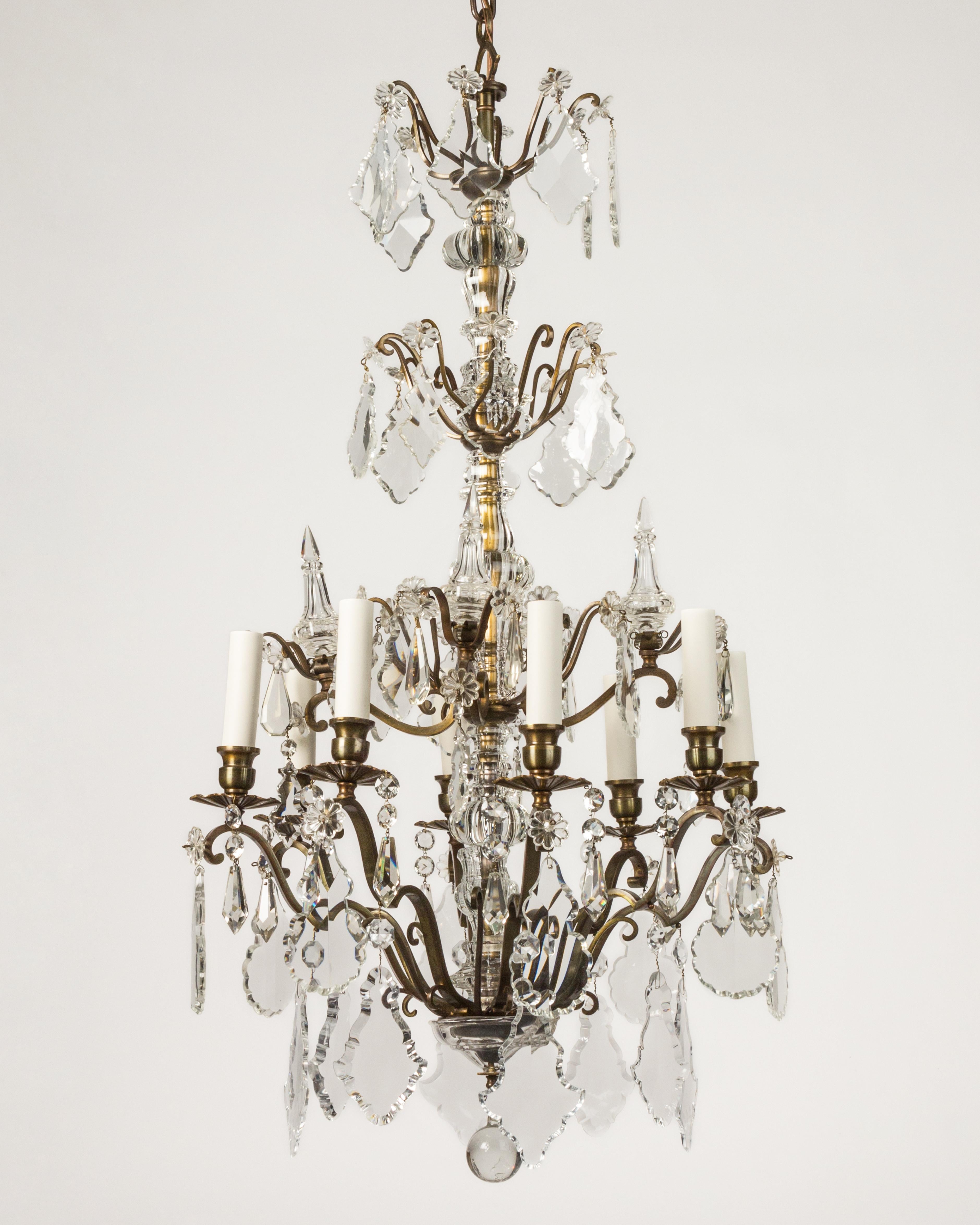 An eight arm chandelier with central glass columns and four tiers dressed with faceted crystal prisms and spires. In its original aged darkened brass finish.

Dimensions:
Current height: 82