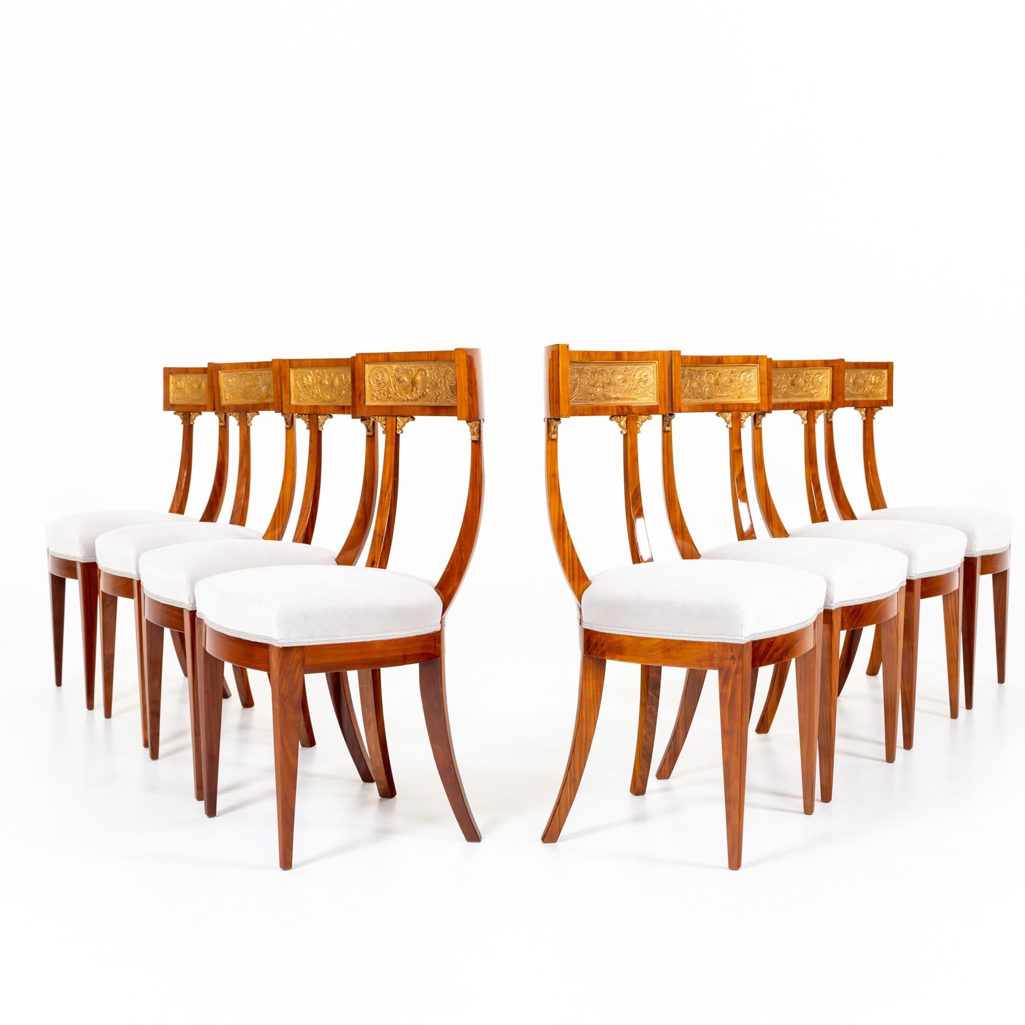 Eight neoclassical dining chairs in the style of antique Klismos chairs with flared and straight tapered legs as well as high backs with curved struts and rounded top part. The upholstered chairs are decorated in the backrest with gold-patinated
