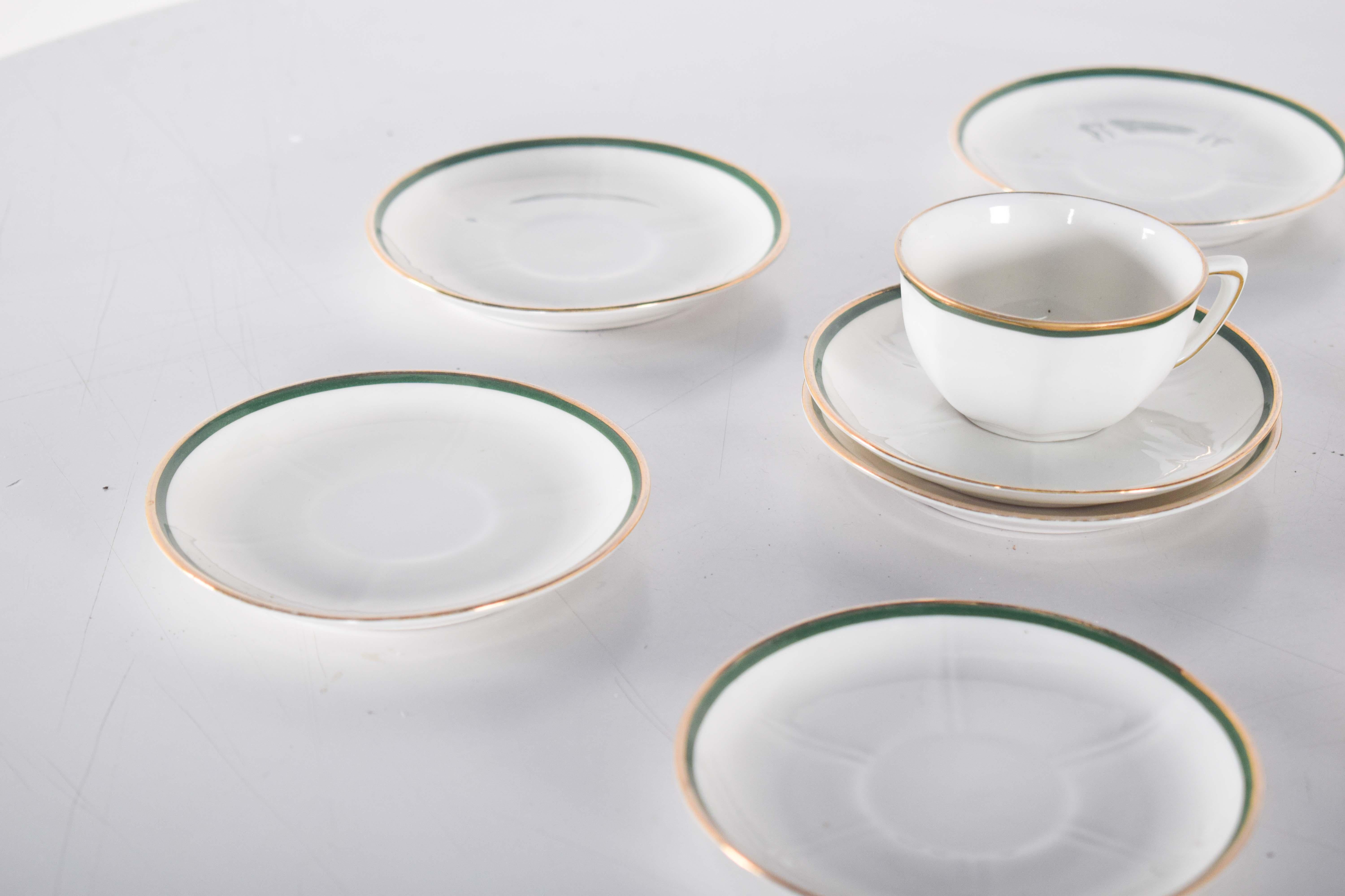 Otto piattini e una tazza in porcellana di Vista Alegre, decorati in verde e oro con un design semplice e classico. Hanno una finitura bianca lucida, messa in risalto da delicate rifiniture dorate sui bordi, che danno un tocco di raffinatezza al