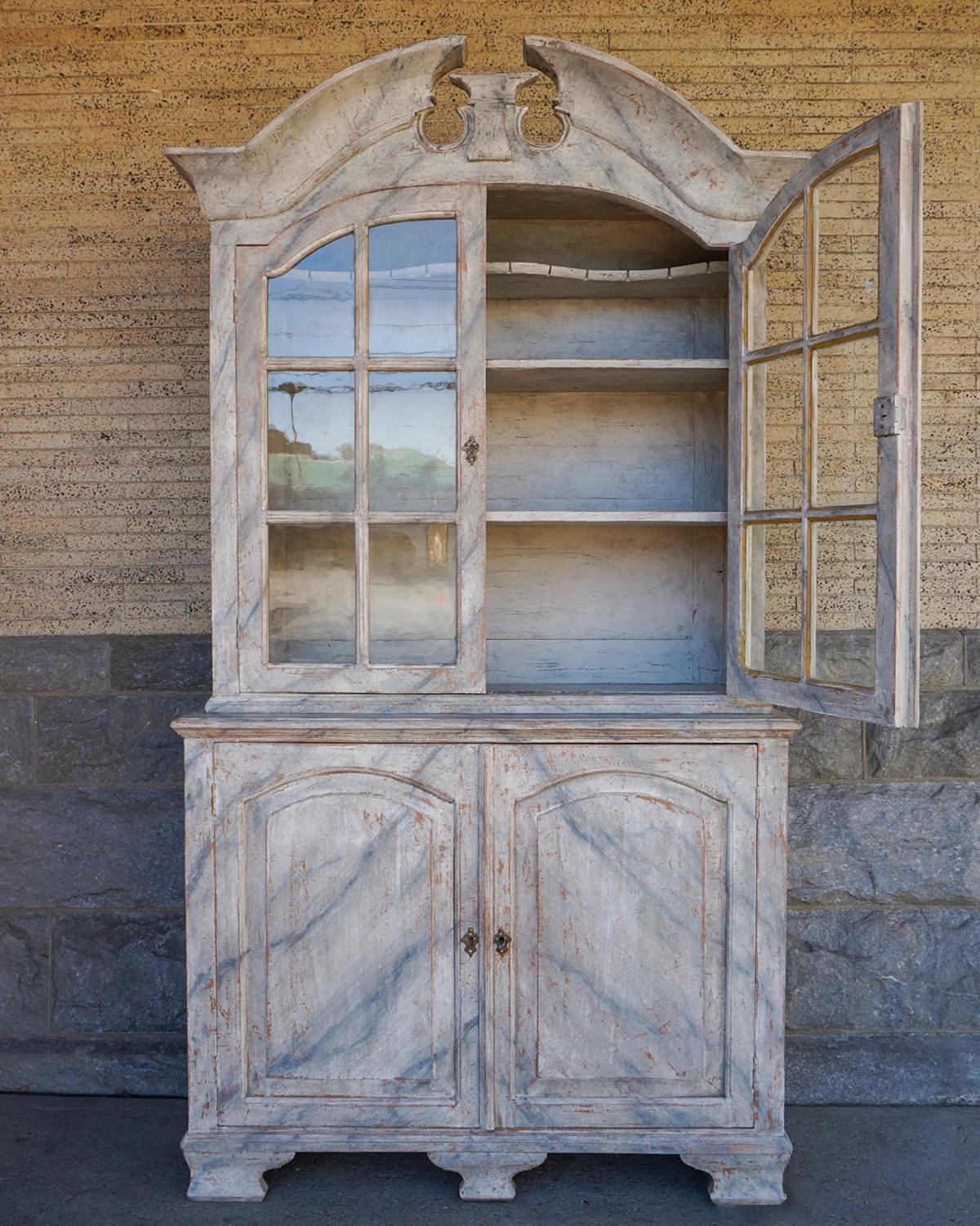 Eighteenth century Swedish cabinet in two parts with overall marbled surface. The upper section has a beautifully carved pediment over a pair of arched doors which retain the original old glass. Inside are three fixed shelves, the uppermost shaped