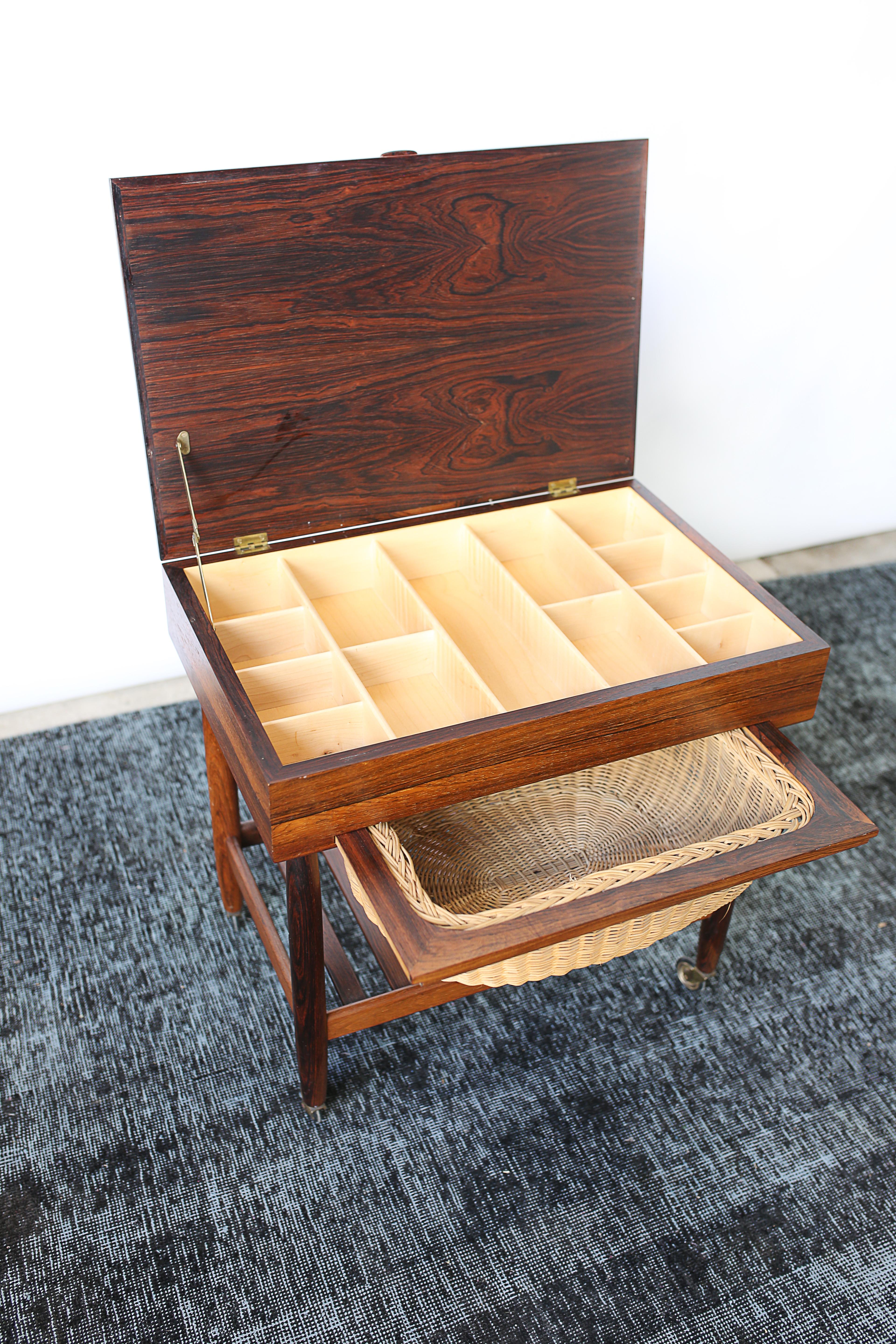 This rosewood sewing table by Ejvind Johannson is in overall good condition. Stylish Danish design. Perfect as a side table. Features include a compartmentalized storage interior, a wicker basket drawer, and a lower shelf with original brass wheel