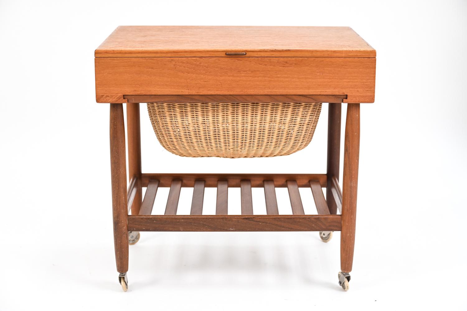 A fabulous Danish mid-century teak sewing cart on casters. Featuring a lift-up top that opens to reveal fitted compartmentalized interior, pull-out wicker basket, and slatted lower tier. On casters. This model was designed by Ejvind Johansson for