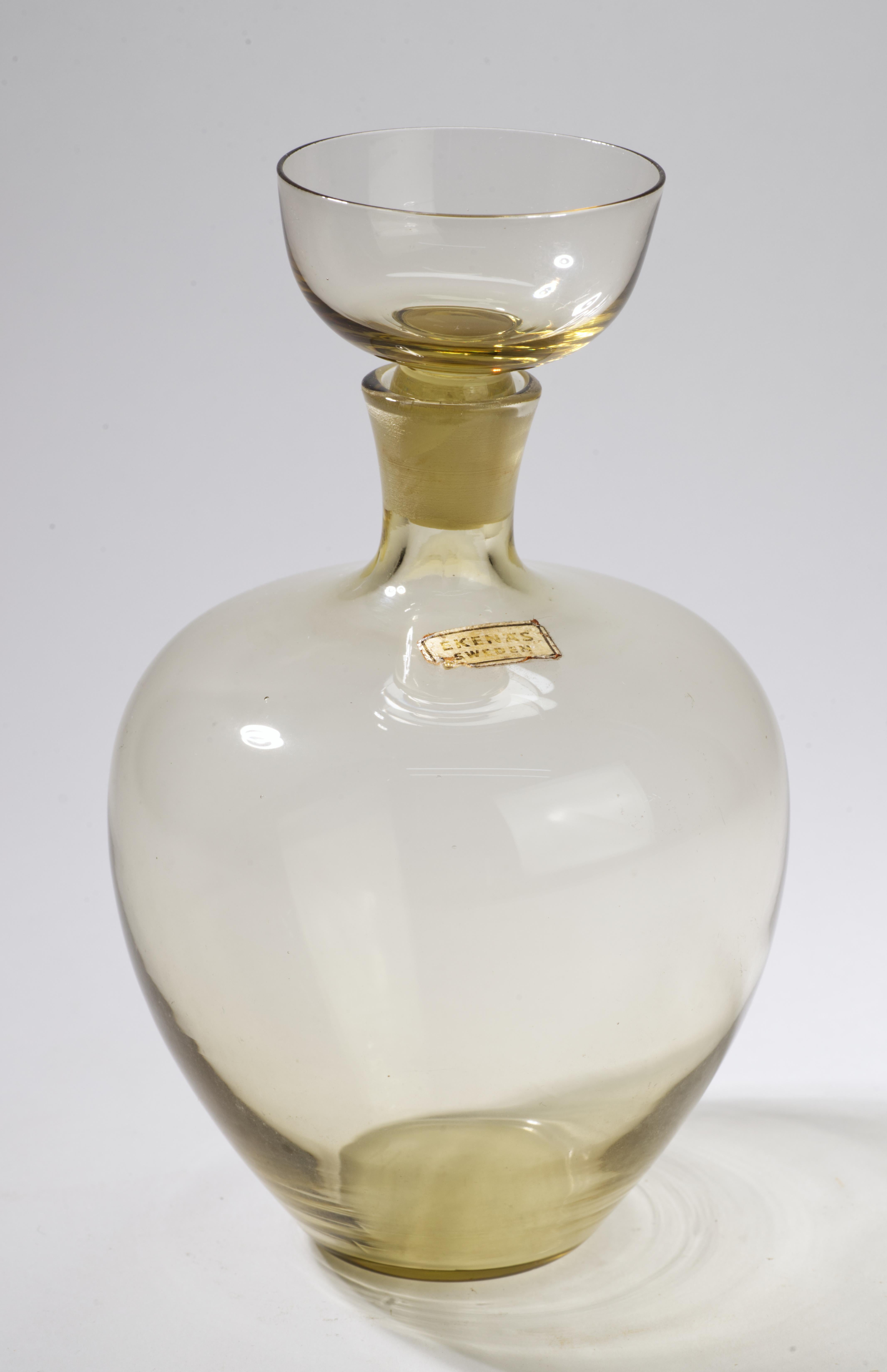  Vintage delicate amber glass decanter has an unusual stopper made in shape of a shot glass. 

The decanter is signed with an original foil label.

Ekenas Glasbruk was founded in Sweden in 1917 by two former Orrefors workers, R. and H. Stahl. In