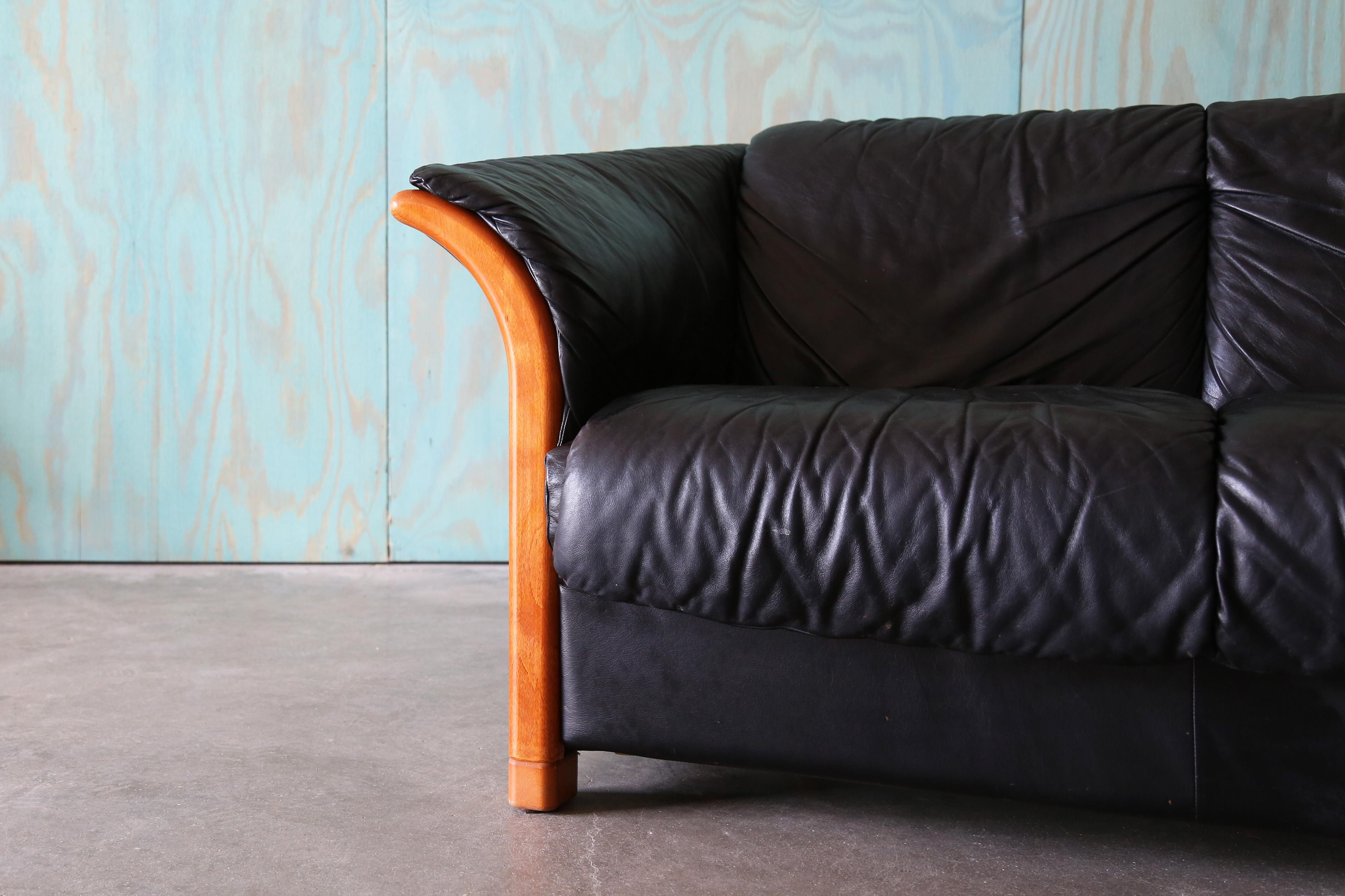 Un Sofá Ekornes Manhattan de piel negra con detalles de madera de teca. Fabricado en Noruega, este sofá está hecho para durar con cuero grueso. Un cómodo sofá de gran diseño con un sutil respaldo curvado y brazos acampanados. Los acentos de teca que