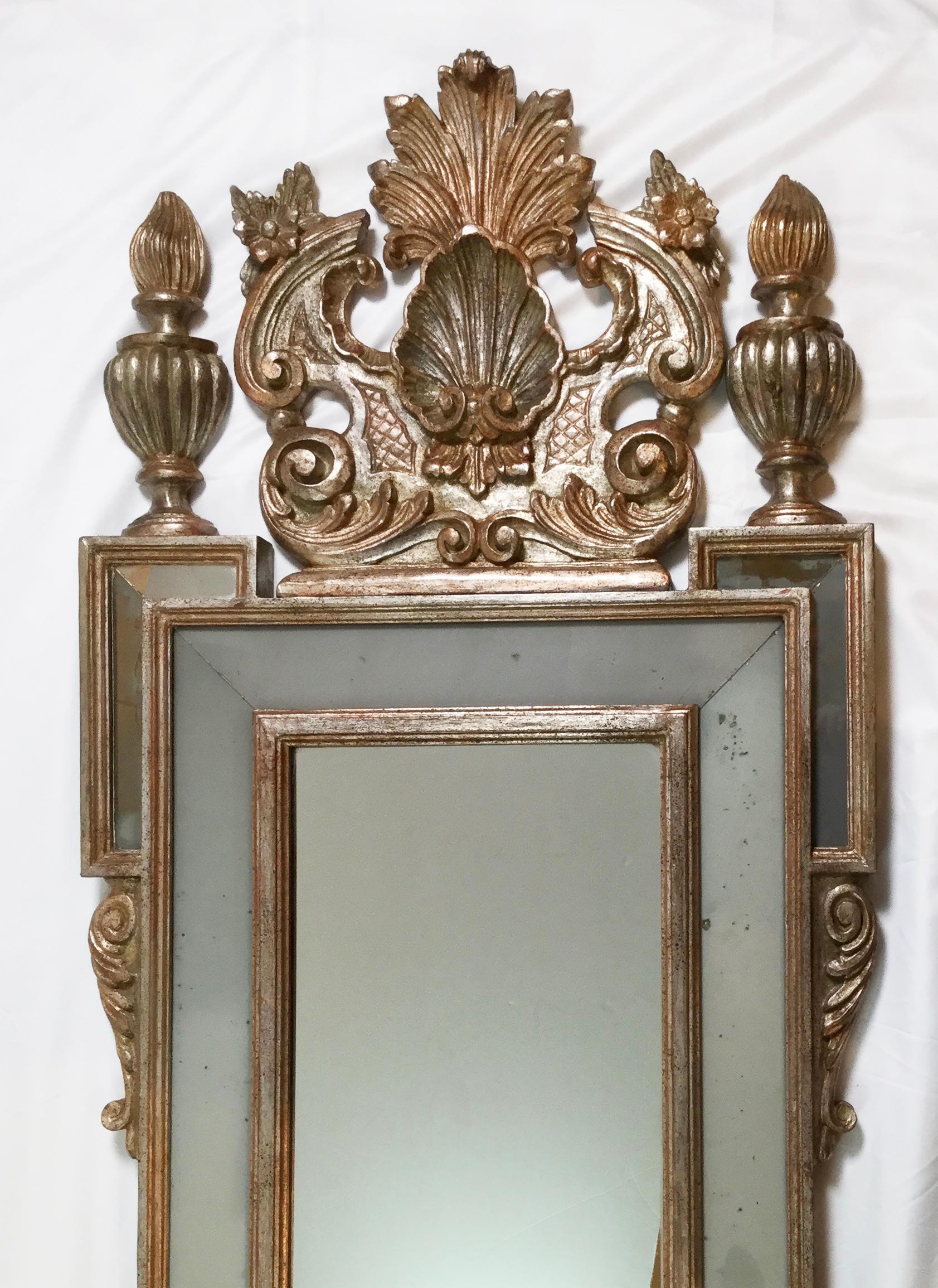Regal silver giltwood mirror with elaborate frame. The top pediment with shell and plume with flanked finials with a soft gray églomisé glass inserts. Beautiful mirror with a slim profile of 25.25 inches and 63 inches tall. The surface is a warm