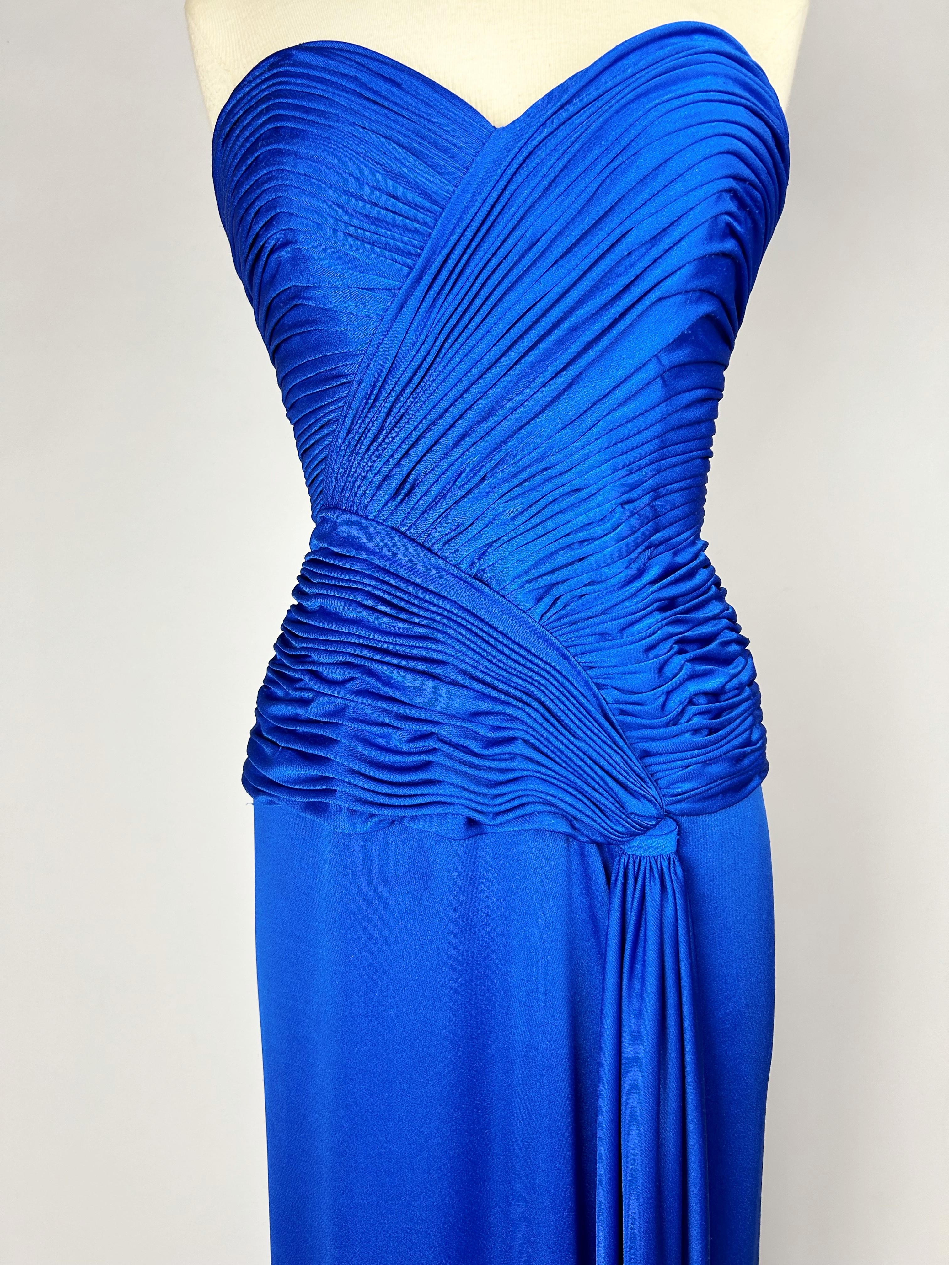 Circa 1980

France

Electric blue lycra jersey evening gown by Loris Azzaro Couture dating from the 1980s. Sheath dress with large plunging neckline and back zip. Bustier with asymmetrical cross pleats underlining the hips and finished with a stole