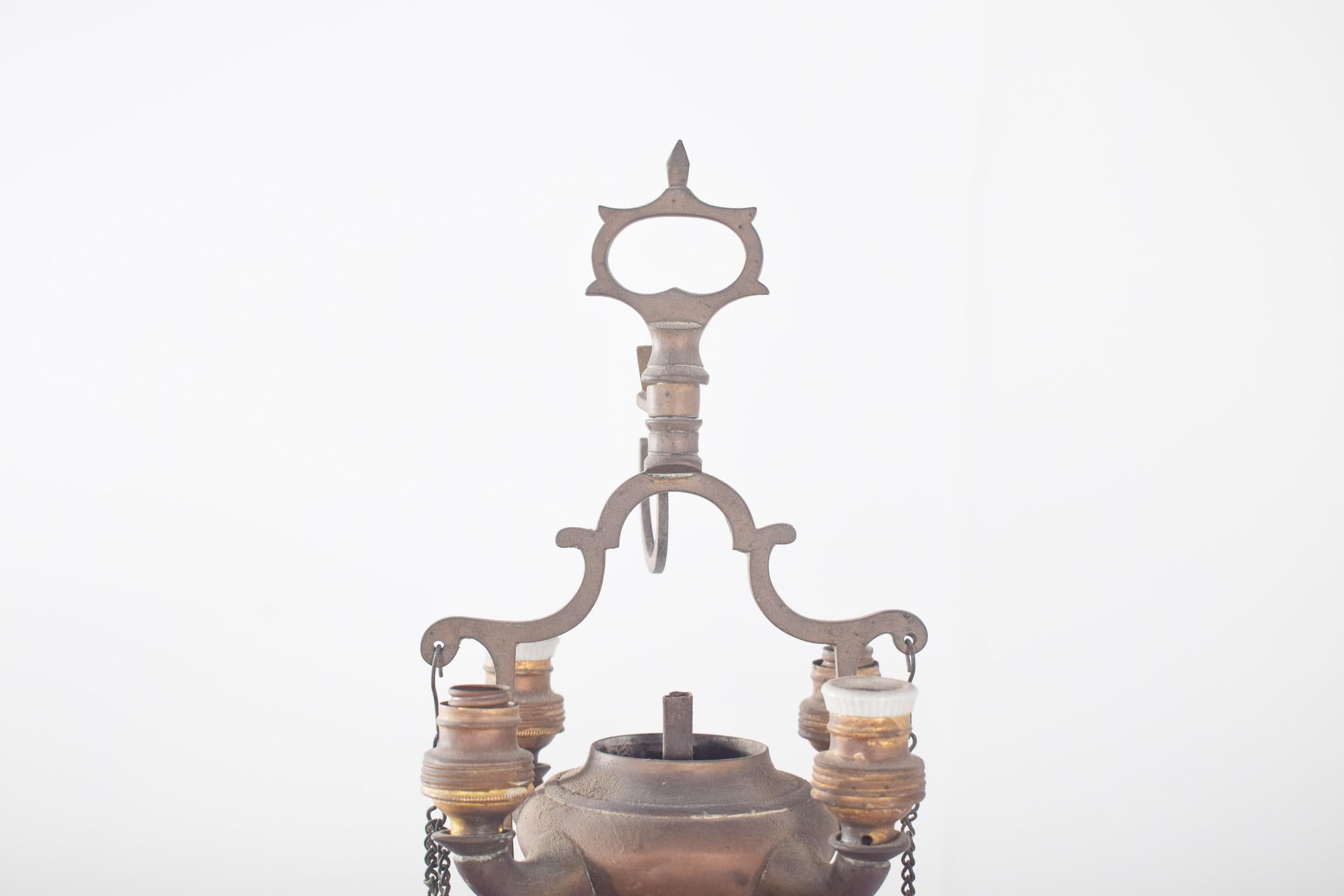 Lámpara de aceite antigua electrificada.  Con una base circular estable de la que se eleva un fuste decorado con detalles torneados. En la parte superior, hay un depósito central para el aceite, con cuatro quemadores laterales donde se colocaban las