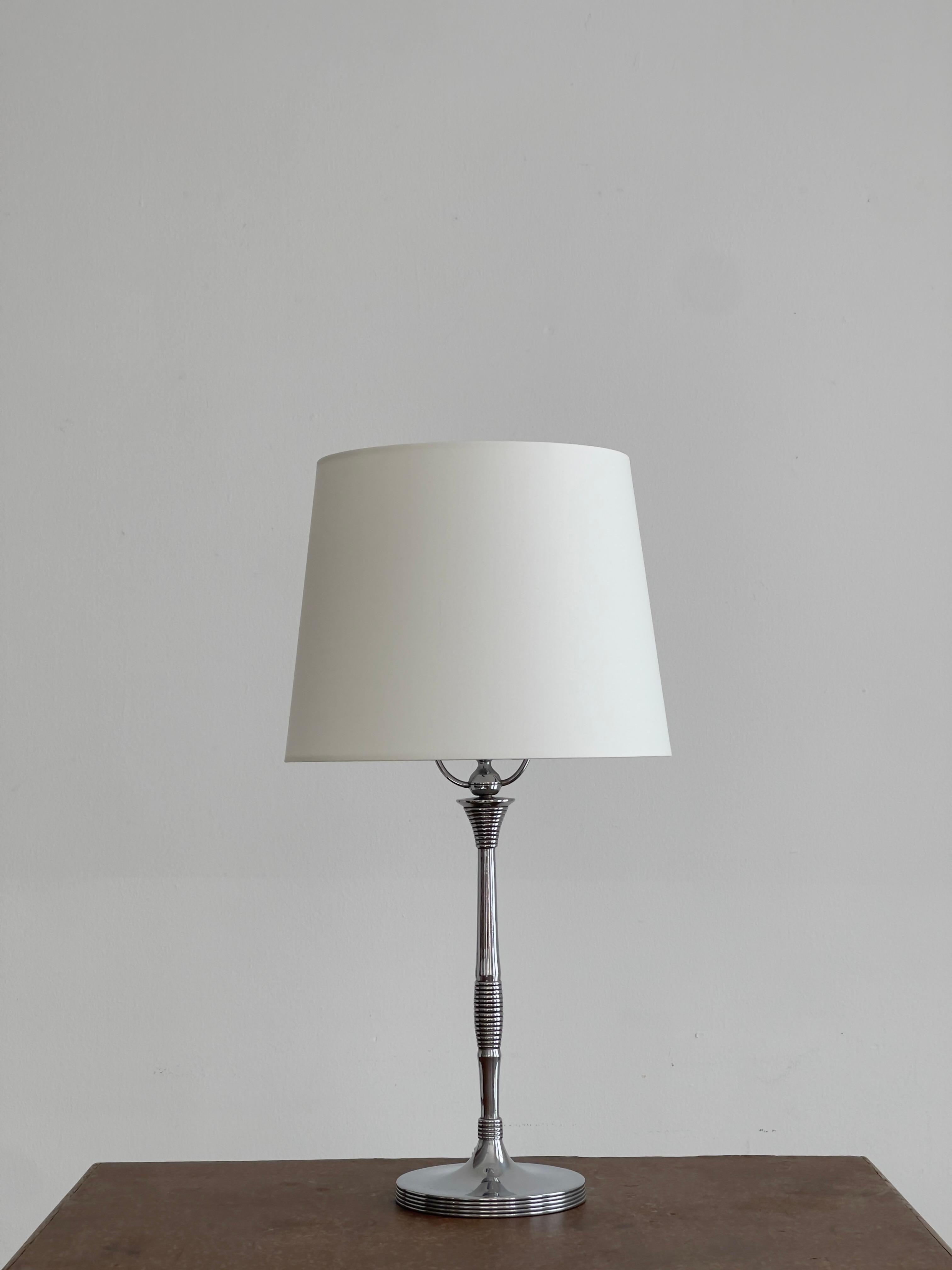 A Danish Art Deco table lamp from the 1920s, composed of nickel-plated metal paired with a porcelain socket, reflecting the decade’s fascination with refined industrial materials. The lamp is distinguished by its layered architectural ornamentation,