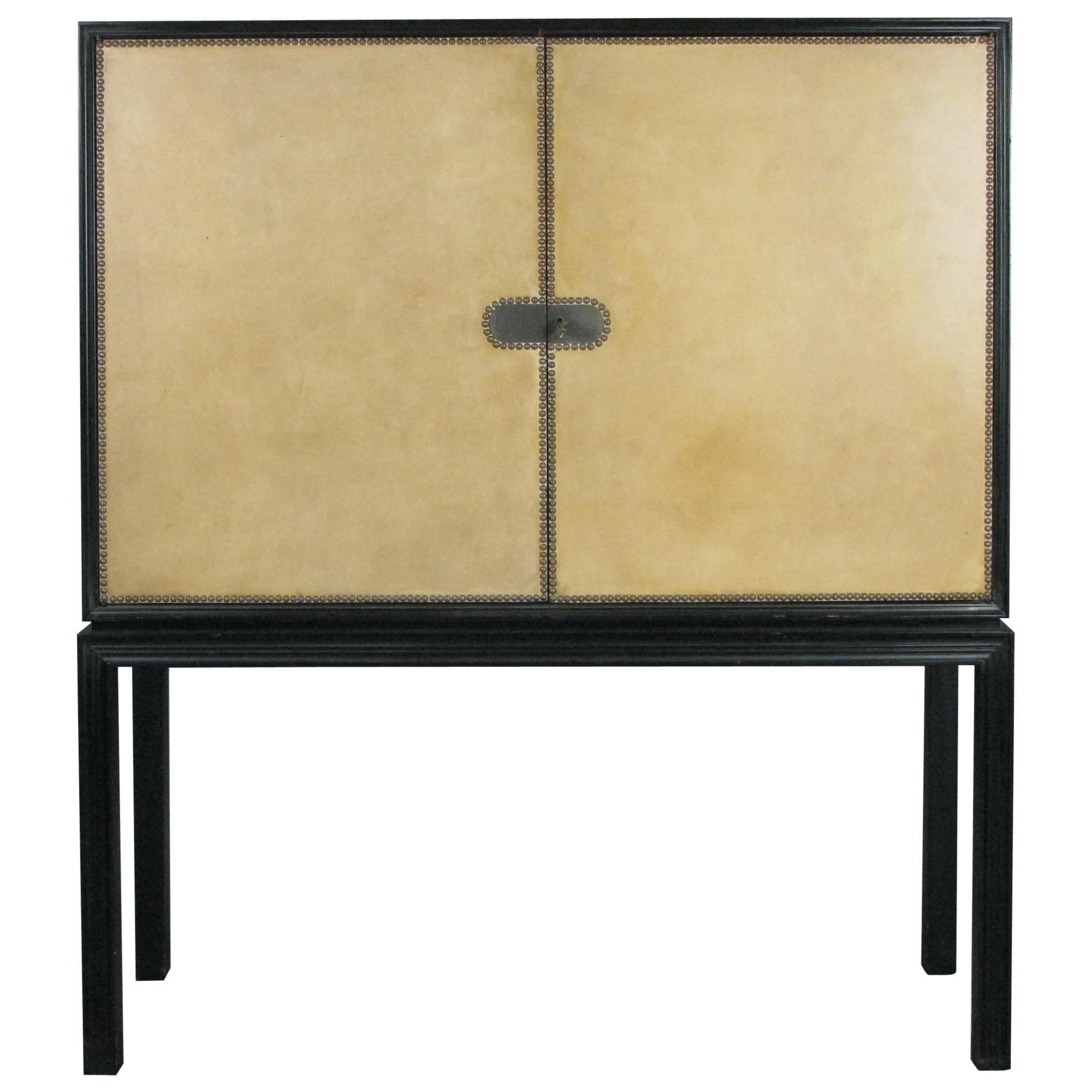 A very elegant and handsome late 1940s lacquered mahogany cabinet designed by Tommi Parzinger for Charak Modern. Raised on tall legs with molded detail, the doors have cream colored leather with nailhead trim detail. The center with bronze plaques