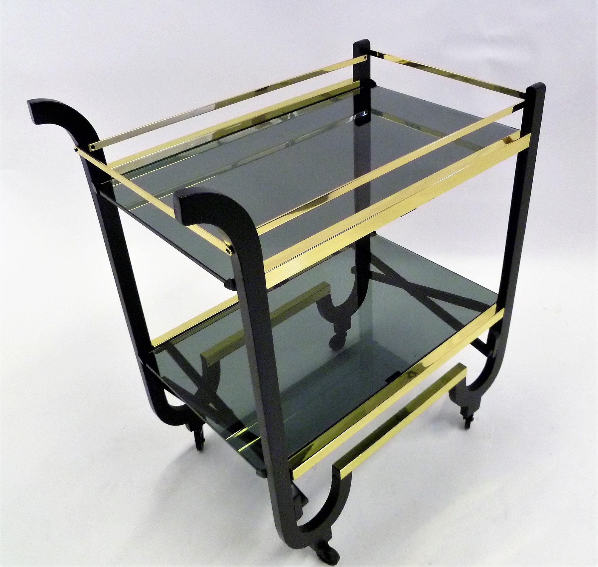 Simply elegant with its brass trims and bars, this 1940s rolling streamline moderne cart features two smoked glass surfaced shelves, Black enameled wood and when the glass shelves are removed, collapsible for storage. Brasses professionally