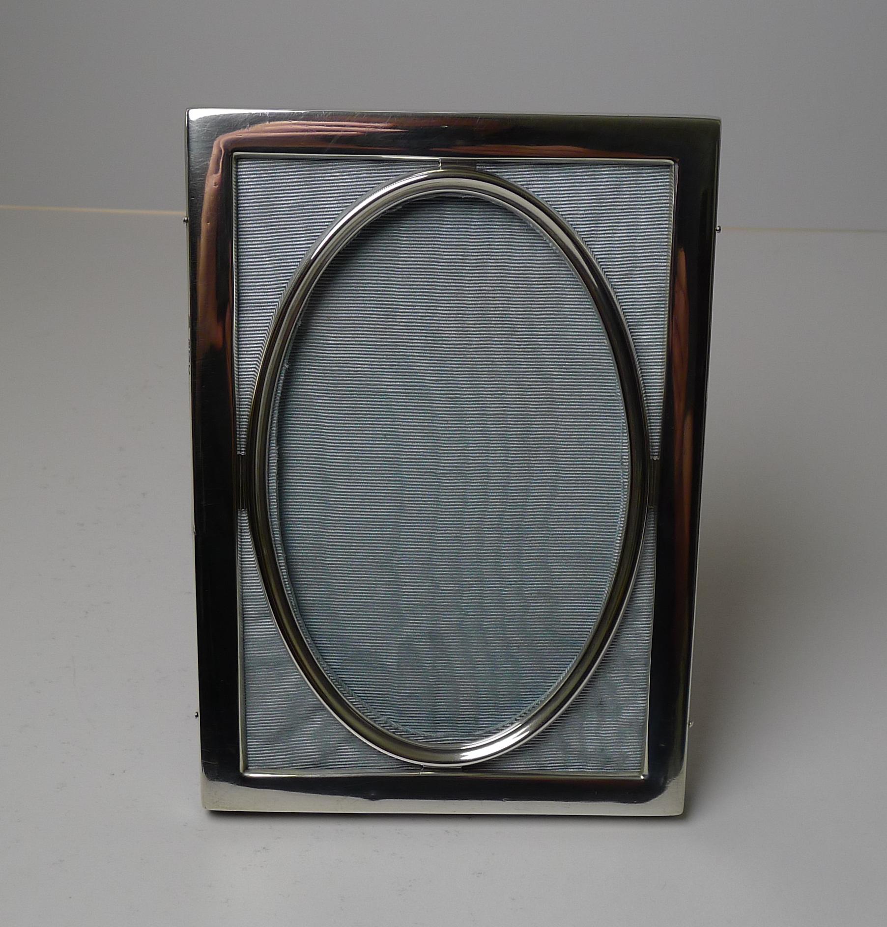 An unusual and very elegant photograph frame made from English sterling silver backed in blue taffeta silk with a glazed central oval aperture.

The back is made from solid English Oak and incorporates a folding easel stand.

The silver is fully