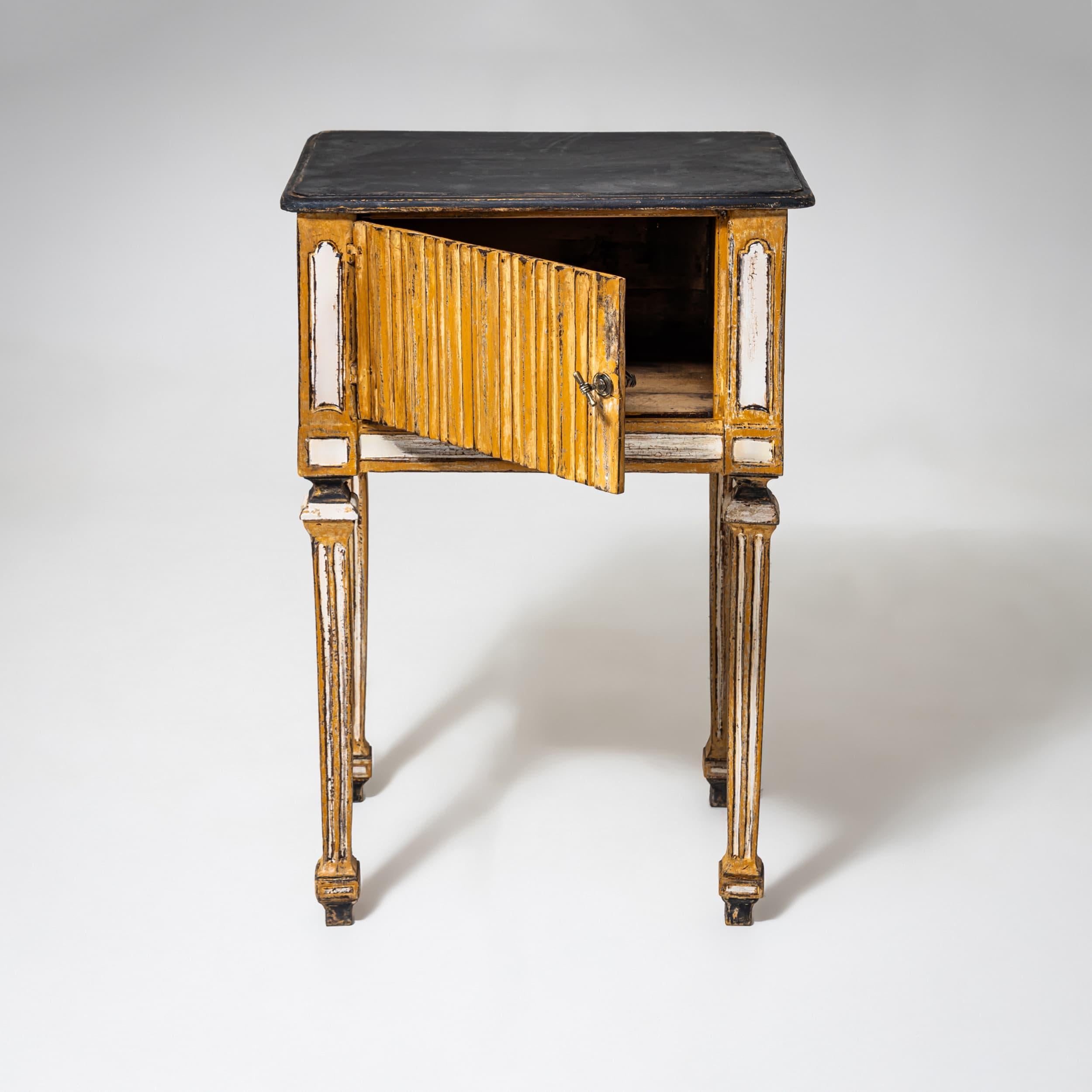 Las esbeltas patas estriadas y los detalles decorativos confieren a la pieza una elegante ligereza. La superficie recién pintada a mano en un cálido tono amarillo con toques blancos y una parte superior oscura resalta su carácter antiguo. La pequeña