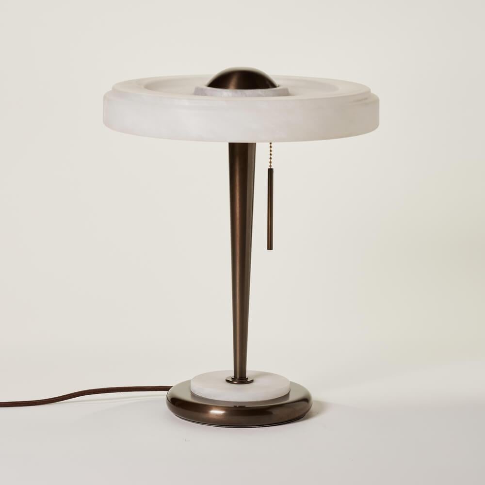 Eine Tischlampe im Retro-Look. Der Lampenschirm aus Alabaster mit einer flachen Form wird von Transparenzen erhellt, die die Maserung dieses Materials betonen. Der Zugkettenschalter erinnert an den Vintage-Stil. Die Kombination aus warmen Farben und