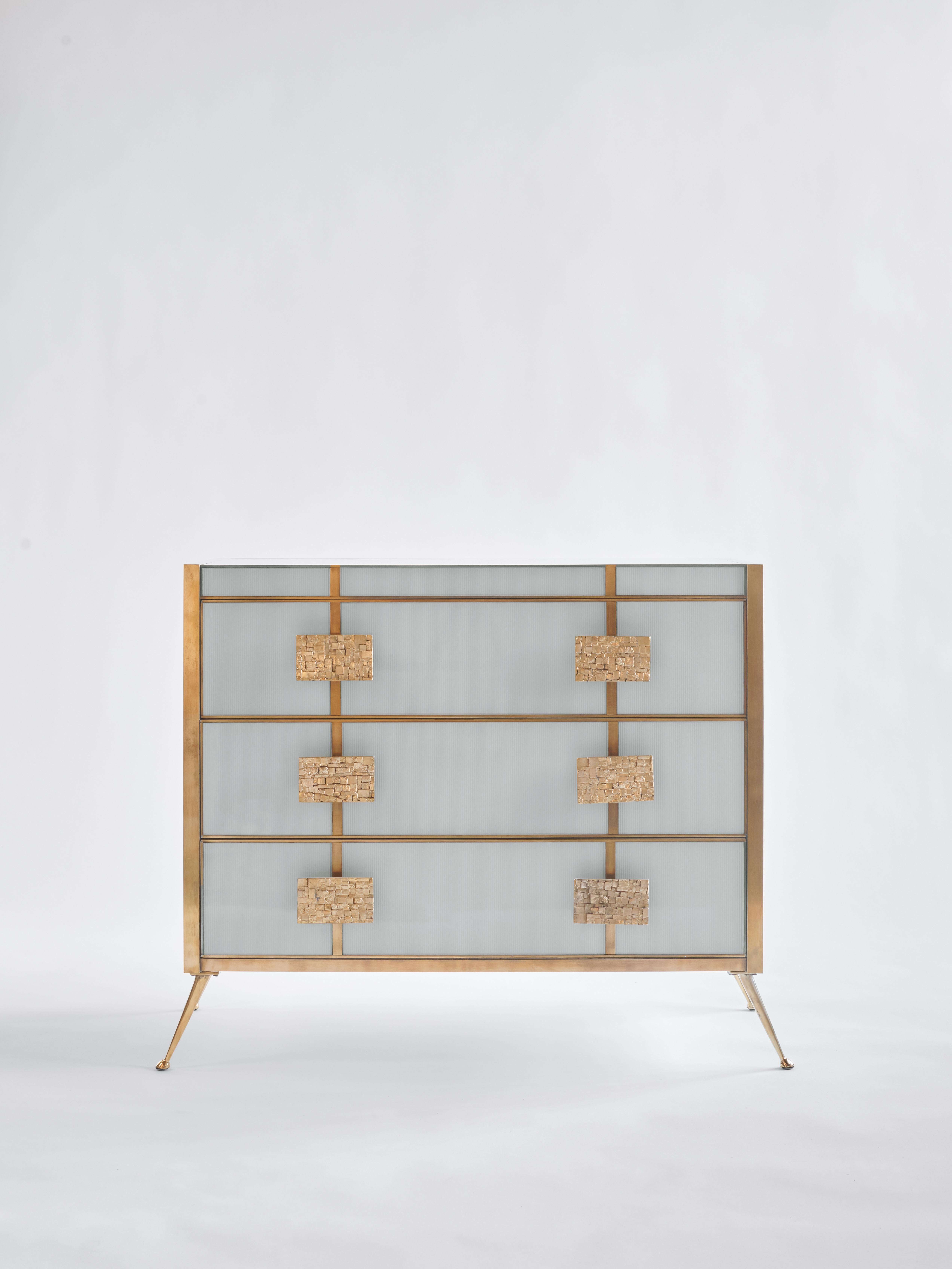 Schublade mit Holzstruktur, verkleidet mit geriffeltem Glas, umrahmt von einem Profil aus gebürstetem Messing, das ihre Geometrie und ihr wesentliches Design hervorhebt. Die Mosaikgriffe aus Messing fügen ein skulpturales und taktiles Element hinzu,