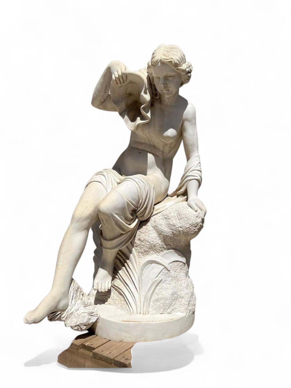 Graceful carved marble fountain featuring a standing female figure holding a shell, evoking classical themes of water and beauty. The sculpture is finely detailed, with flowing drapery and soft facial features, carved from solid marble with refined