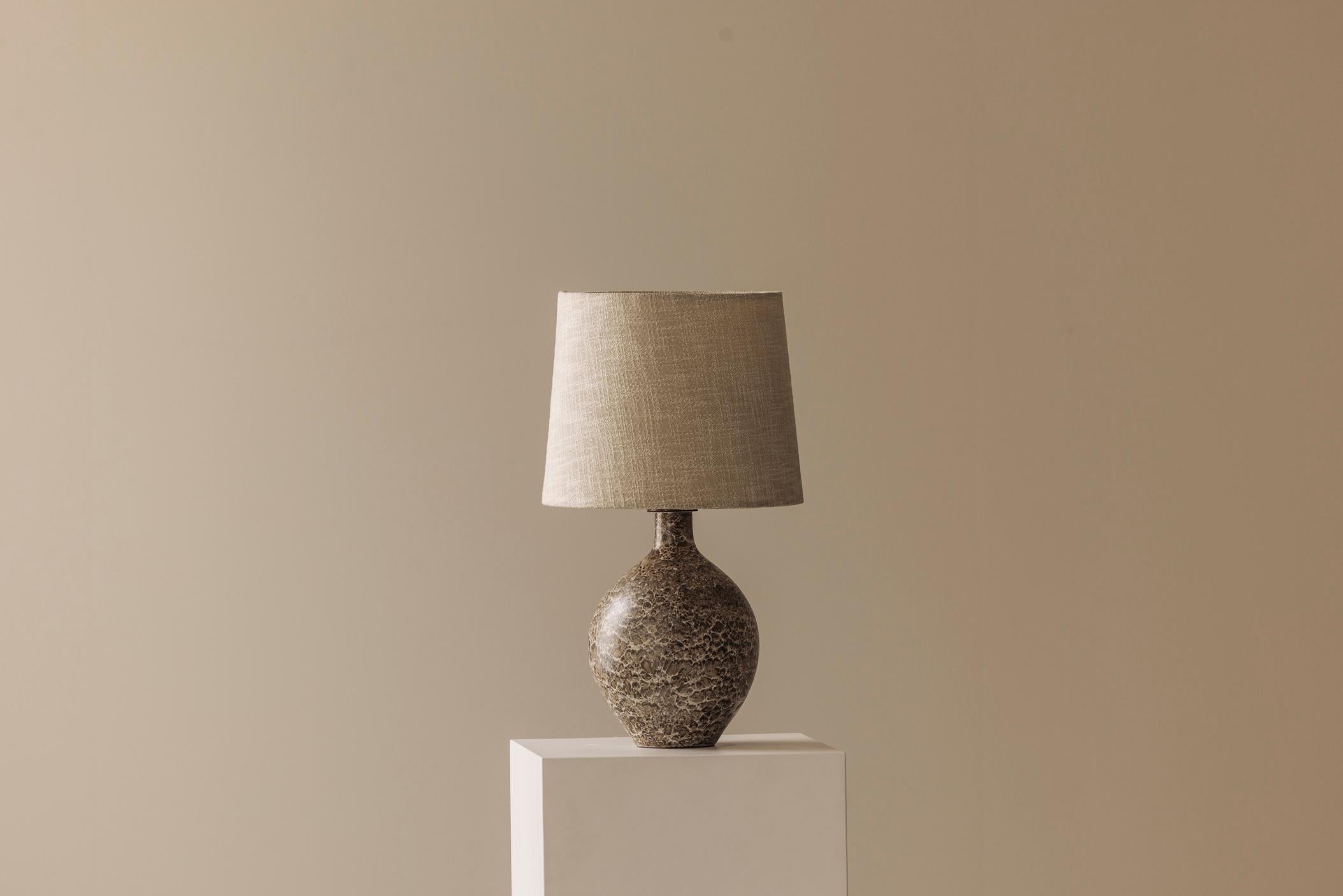 Admire this elegant ceramic table lamp sourced in France dating from the 1960s. It features a richly textured ceramic base in warm, natural tones. The lamp is fitted with a new linen shade that adds a clean, modern finish while respecting its