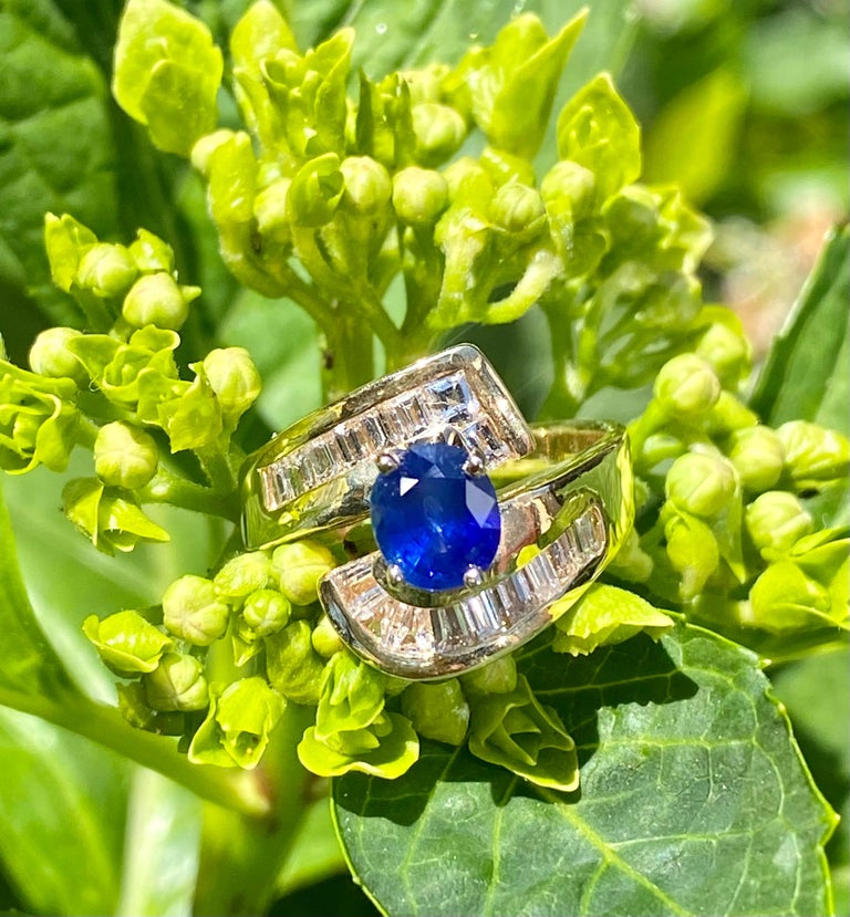 Classic bypass style 14 karat yellow gold estate ring features a vivid, cornflower blue oval shaped natural sapphire, prong set with 4 prongs, and surrounded by approximately 1.75 carats of channel set square and tapered baguette diamonds.  Sapphire