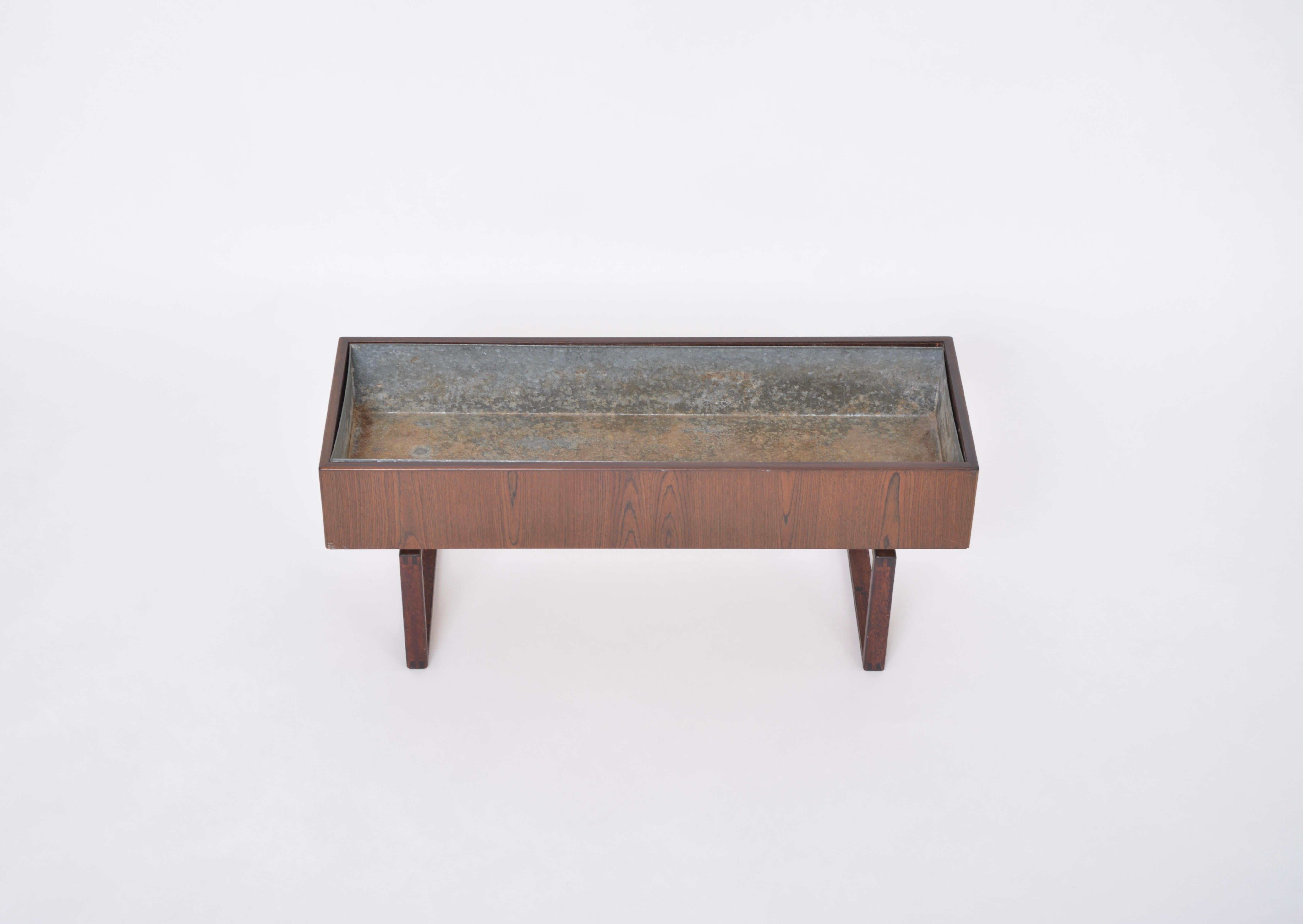 Questa fioriera per interni è stata prodotta negli anni '60 in Danimarca. Con un design molto grafico e minimale, la fioriera poggia su due gambe di forma quadrata. La fioriera è realizzata in teak con una vasca in metallo intarsiato. Ottimamente