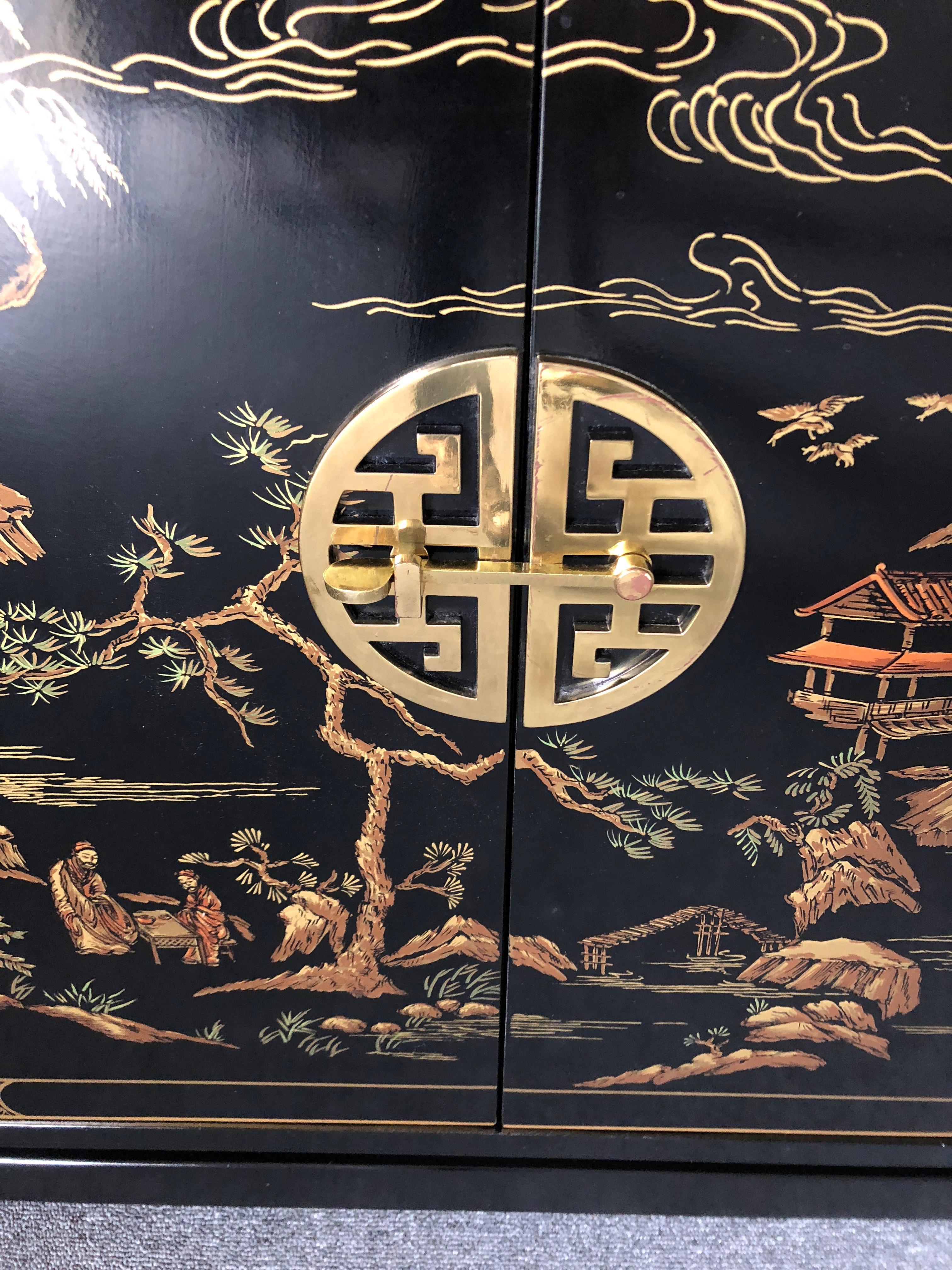 Elegant sleek black chinoiserie two-door cabinet having lovely scenes of Asian landscapes and figures in gold, cream, and orange, with a handsome round Chinese style latch and single shelf inside.