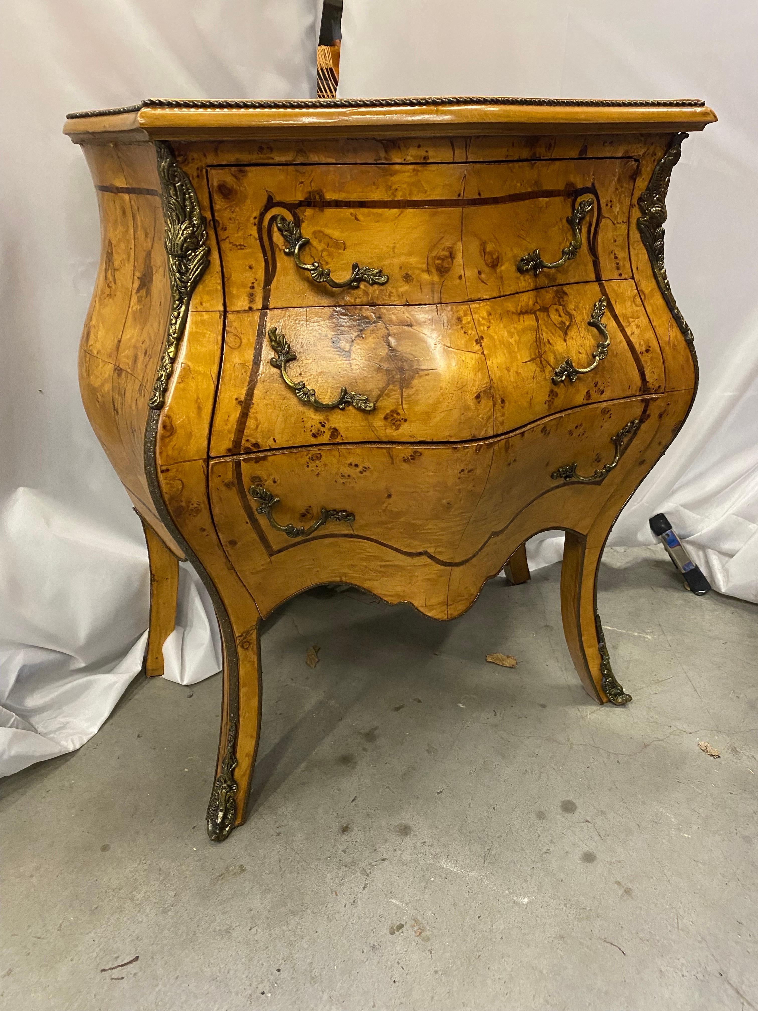 A very stylish diminutive bombe chair or bed side chest.  The highly figurative book matched wood with cast brass mounts.  The light olive wood with a darker inlay around the top, sides and drawer fronts, the finish has been professionally buffed