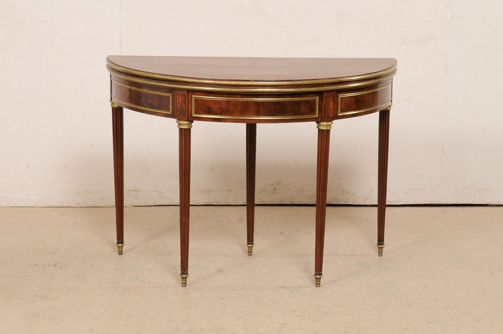 An elegant French Neoclassical demi-to-round table with brass accents from the early to mid 19th century. This antique demi-lune table from France features a half moon top over a rounded apron, and is raised upon five flute-carved rounded legs, that