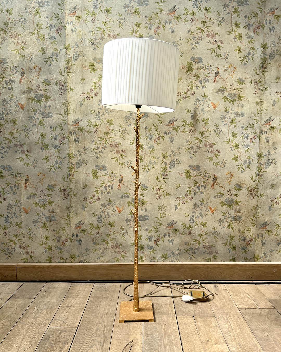 A refined floor lamp with a slender, textured hammered metal stem finished in a rich, luminous gold. The branch-like design adds a sculptural presence, while the pleated white lampshade diffuses a soft, warm light. The lamp sits on a square metal