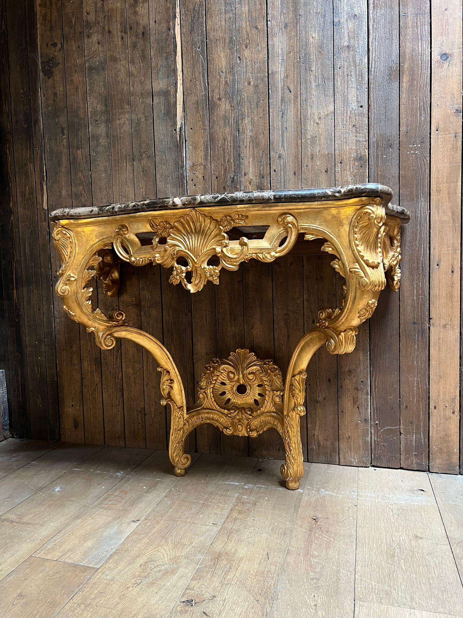 Elégante console en bois doré, richement sculptée dans le style Louis XV, surmontée d'un beau marbre brun veiné. La console est ornée de rinceaux, de guirlandes florales et d'un motif central en forme de coquillage, le tout mis en valeur par une