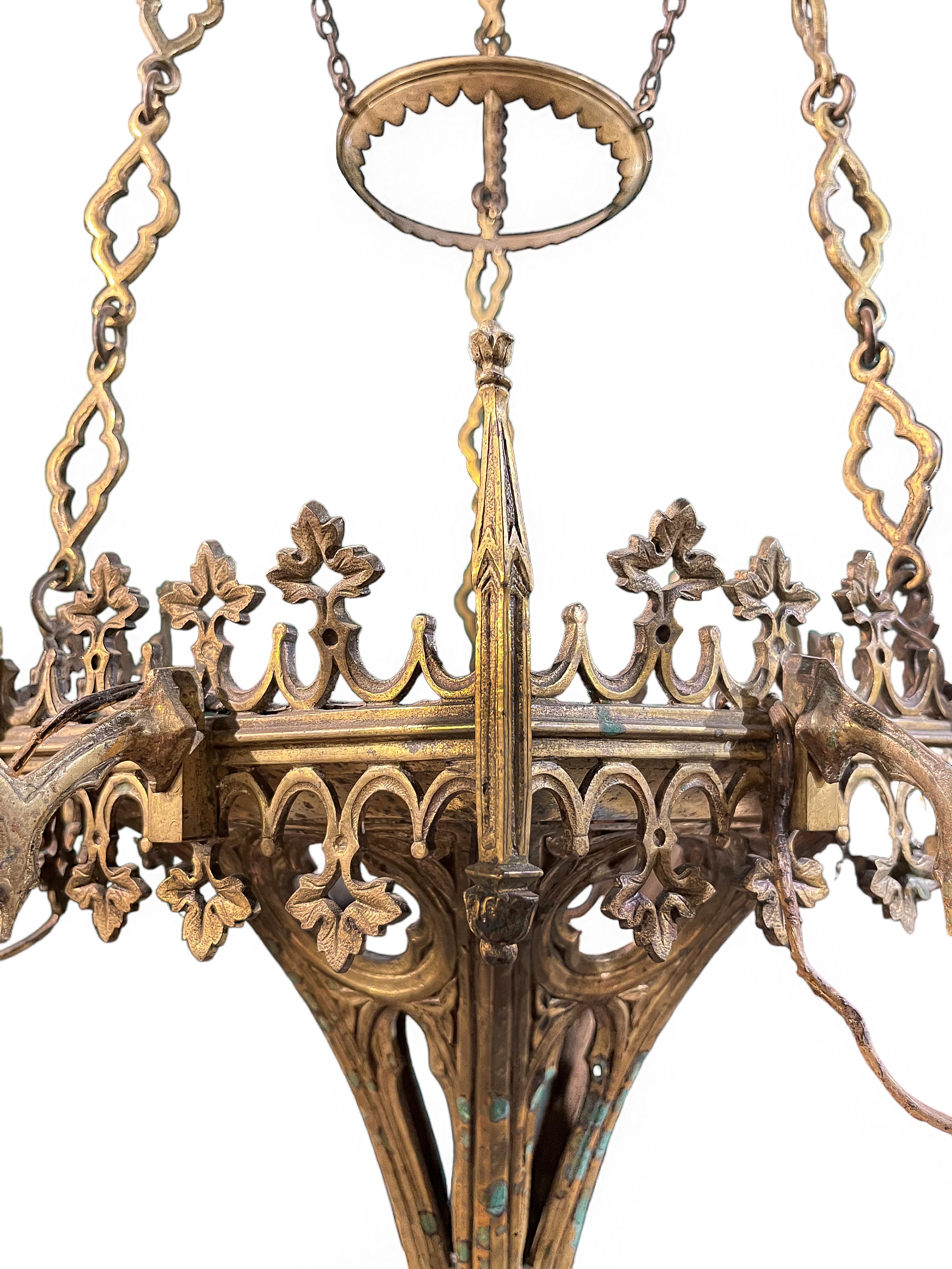 The bronze chandelier is comprised of a foliate canopy with elongated quatrefoil link chains, suspending a tapering basket encircled by gothic grape vine tracery and mounted with six arched candle arms. The basket is cast with quatrefoils above