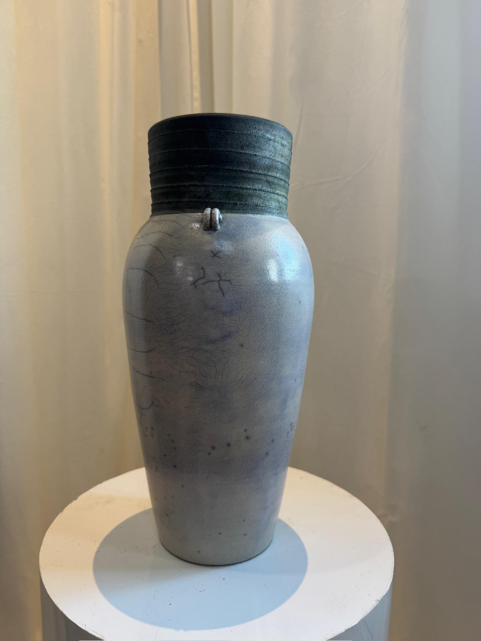 Esempio di ceramica Raku, questo vaso in ceramica lavorato a mano presenta un caratteristico smalto crepla che ne esalta l'aspetto organico e invecchiato. La forma elegantemente affusolata è accentuata da un bordo scurito a coste, che crea un