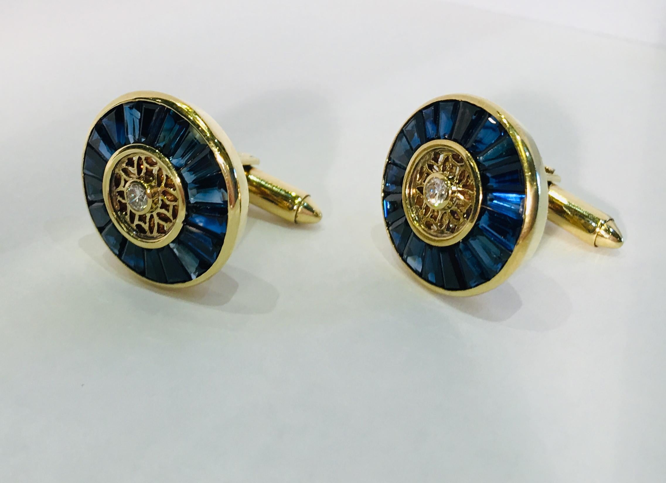 Impeccably styled, very unique 18 karat yellow gold estate cuff links feature vivid blue sapphire baguettes, channel set in a circle, with bezel set round brilliant diamond centers, separated by patterned, openwork fretwork gold. Sapphire is the