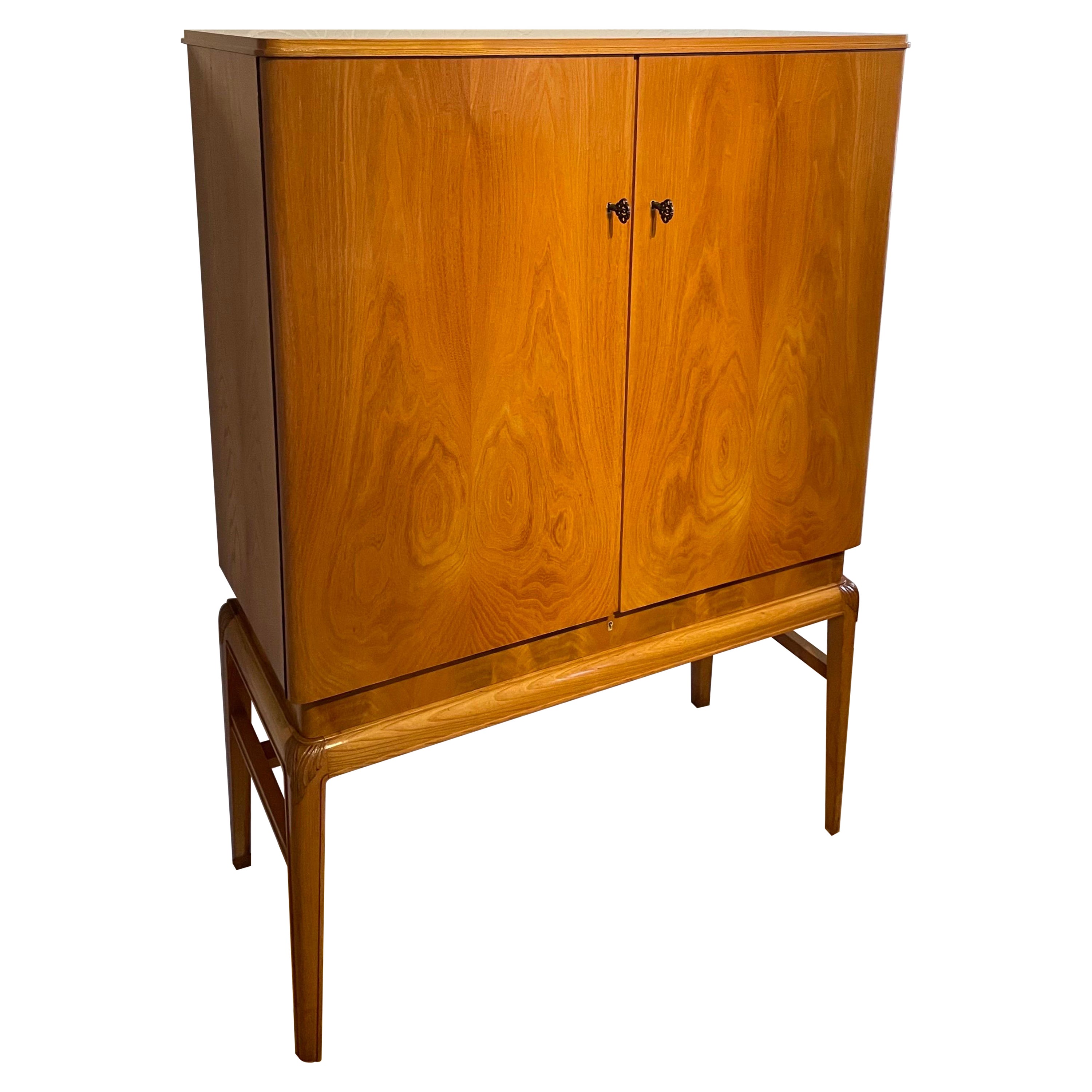 This is the 1930s Swedish Grace cabinet made from elm and birch in nearly excellent condition.

It comes with a tall elegant leg structure with soft rounded “hips” decorated with hand carved ornaments.
The tapered mid section gives the cabinet a