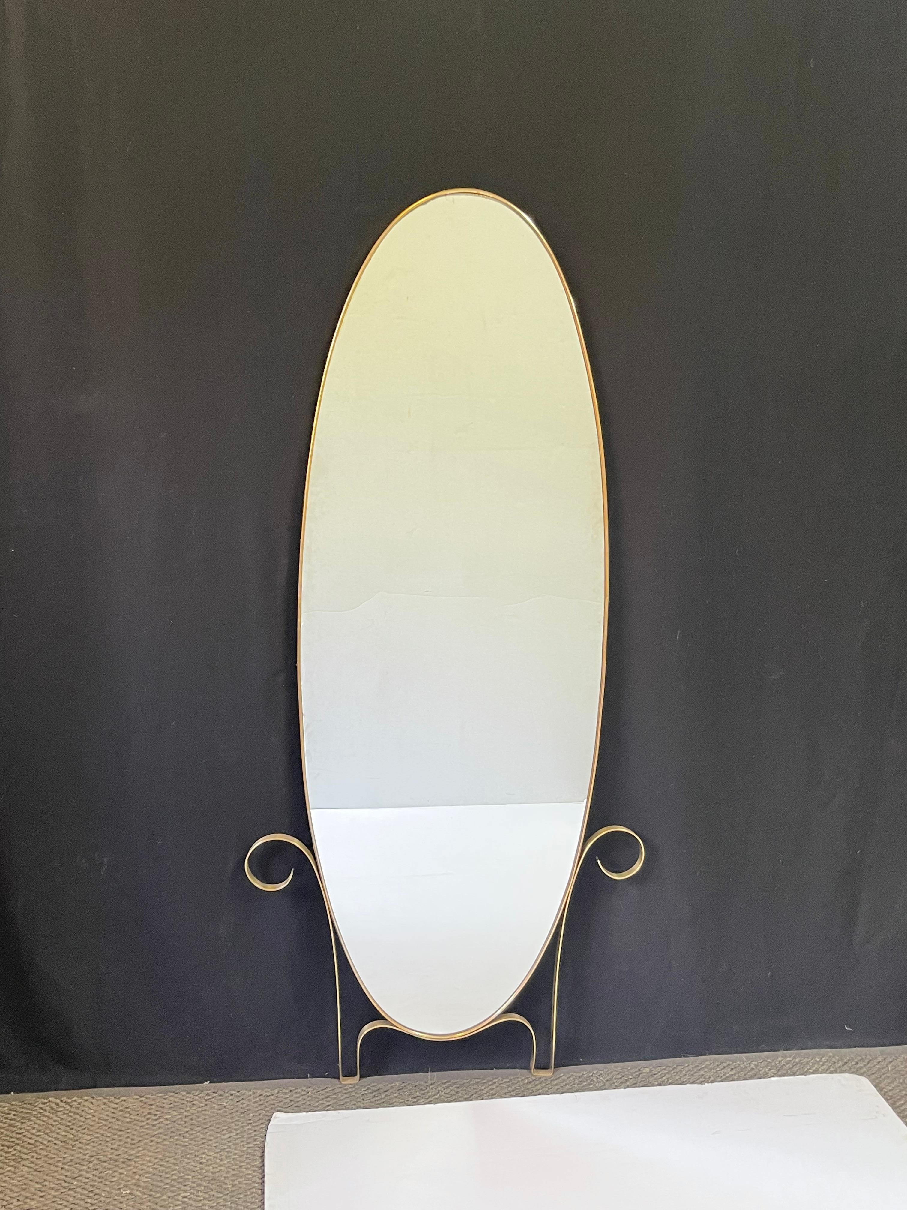 Large mid 20th Century modernist Italian elegantly elongated and tapered wall mirror with a slender oval brass frame. The mirror features a brass detail at its base with a playful upswept scroll. The original mirror-glass and brass frame are in good