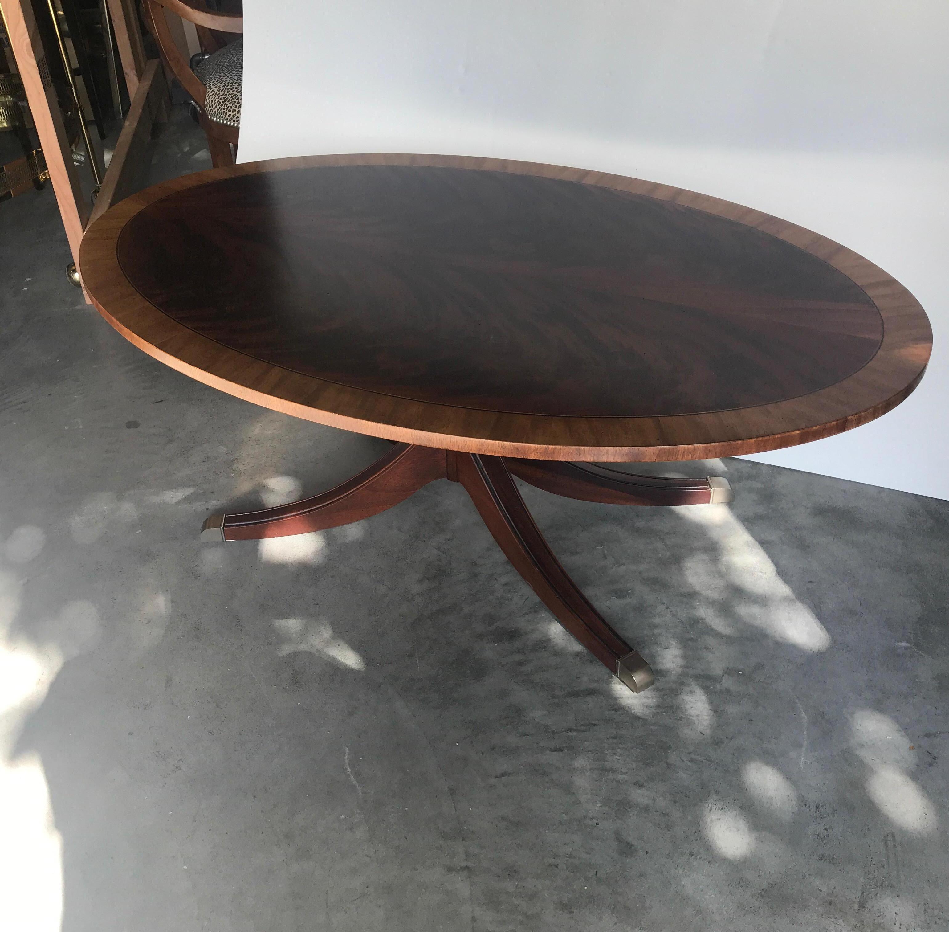 Graceful oval flame mahogany cocktail table with pearl wood border. The generous size top with central column and classic Duncan Phyfe four leg base and brass capped feet.