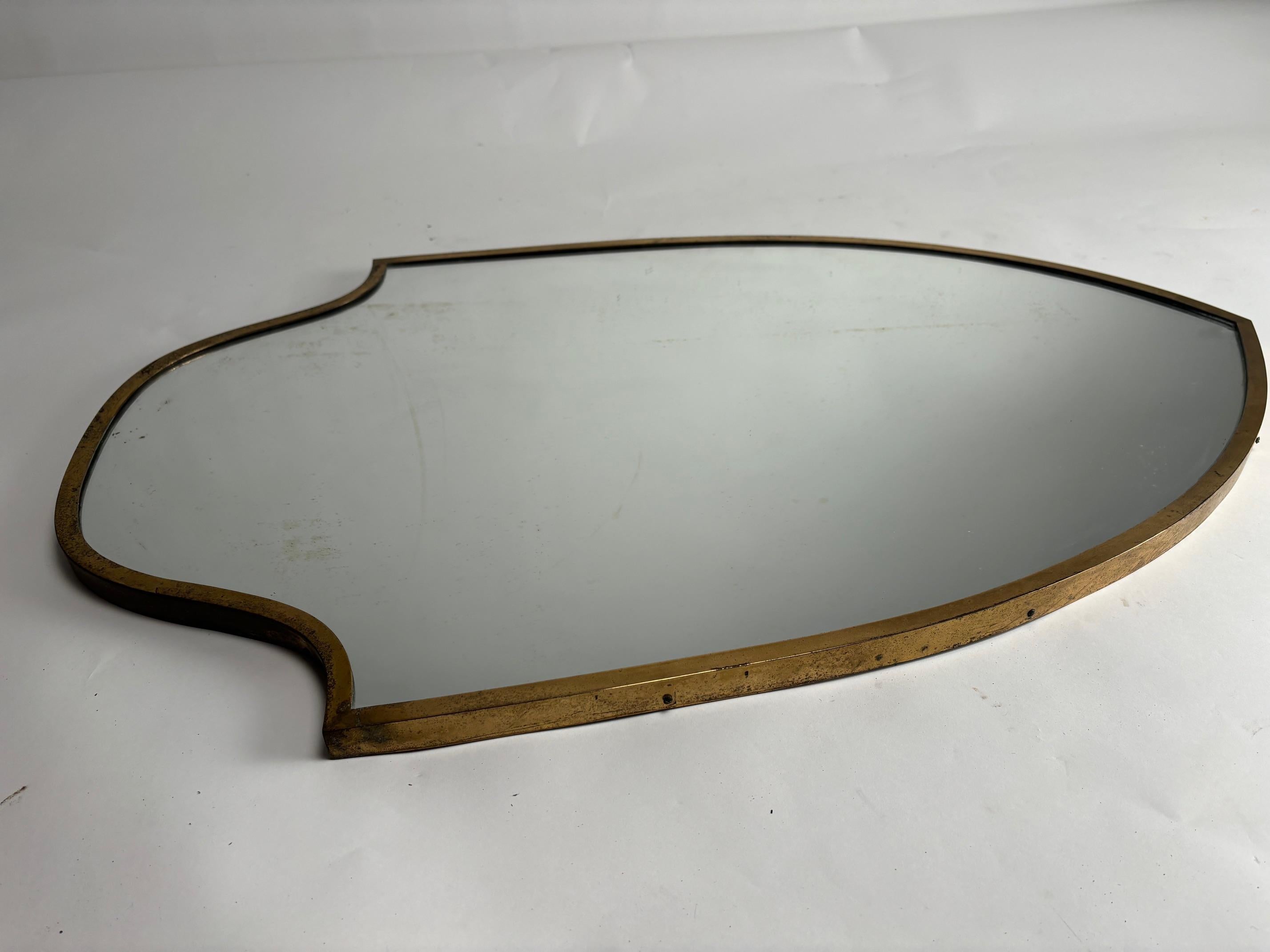 This mid-century brass mirror recalls Gio Ponti's elegant and refined style with its sinuous, sculpted frame, evoking a sense of movement and harmony typical of Italian design of the period. 
The shield-like shape, with rounded edges and a slightly