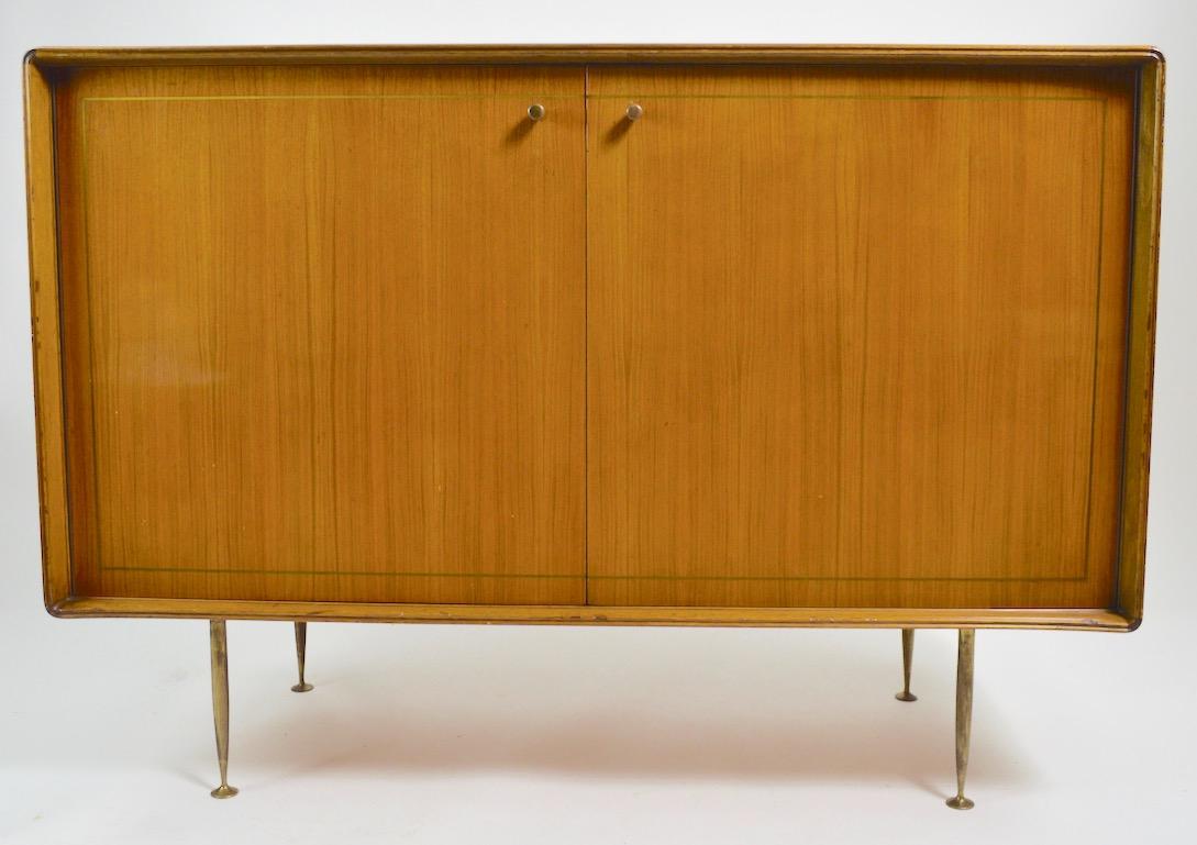 Chic and stylish two-door cabinet which opens to interior drawers, as shown. The case has shaped sculptural edges, tapered brass legs, brass pulls, and inlay brass trim. It is in original finish, which shows cosmetic wear normal and consistent with
