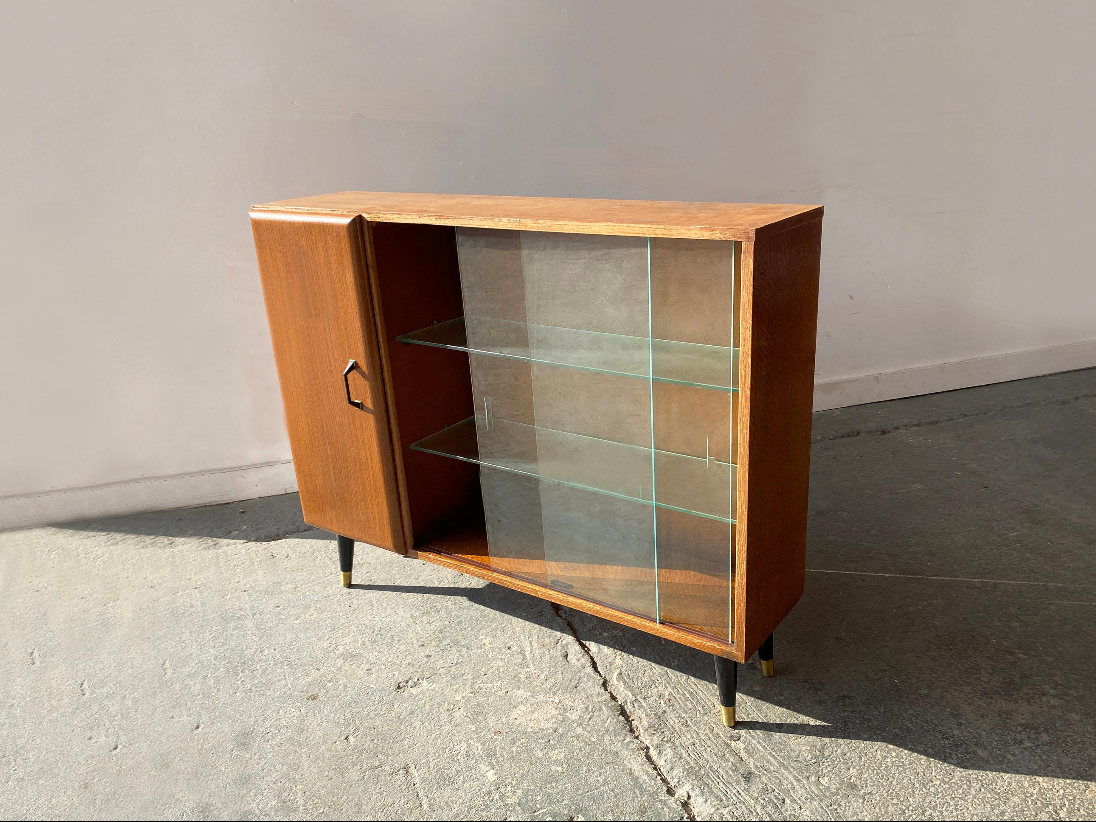 Une bibliothèque en teck avec une seule armoire. Les deux étagères en verre sont bordées de portes coulissantes en verre. La bibliothèque présente des détails de design subtils : une poignée de porte en métal noir et laiton et des pieds assortis en