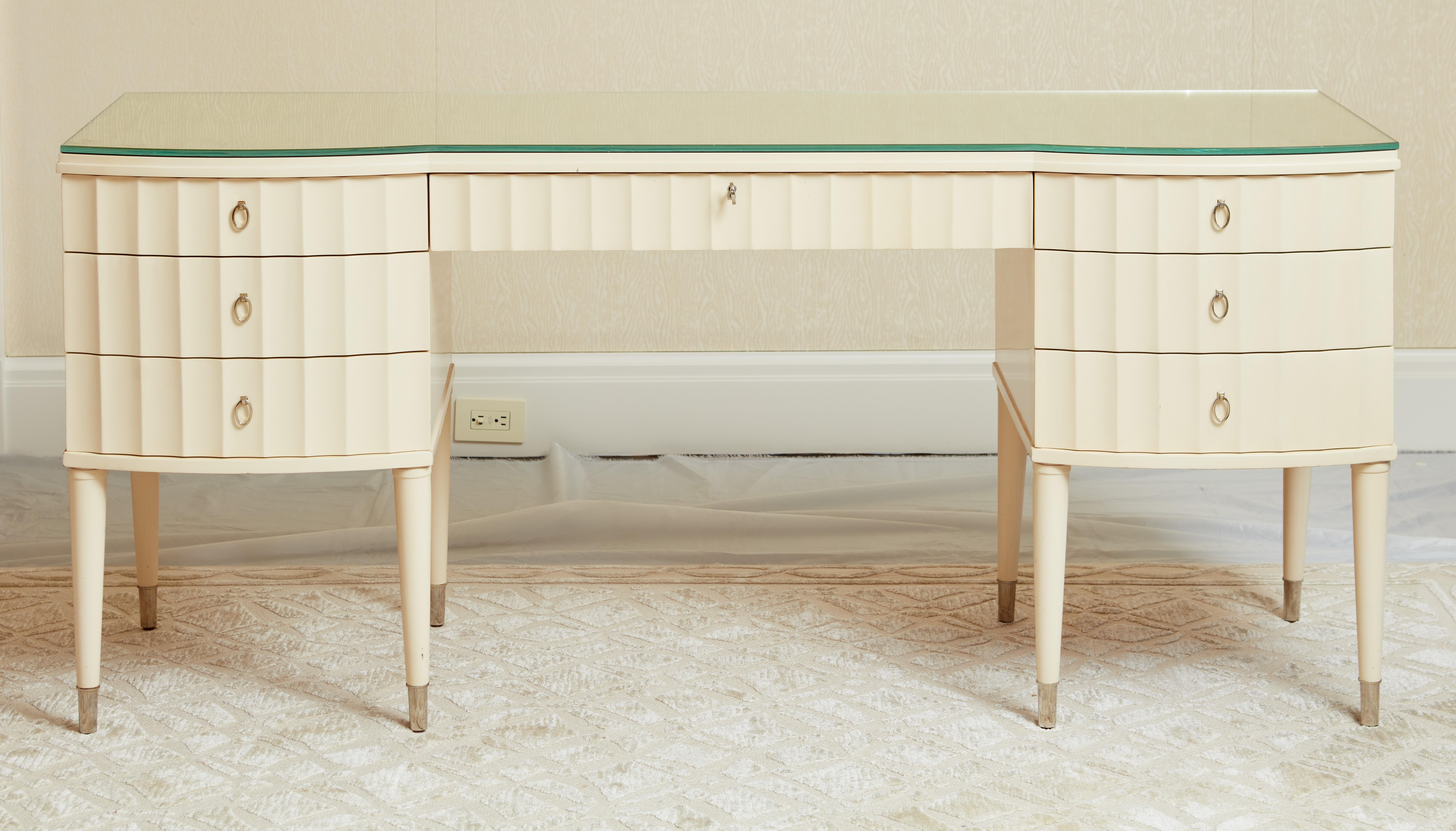 The cream lacquered dressing table having an antiqued mirrored topresting on a cream-colored dressing table / vanity. The front with a fluted surface with a central drawer flanked by 3 sets of drawers on each side, all supported by 8 turned legs on