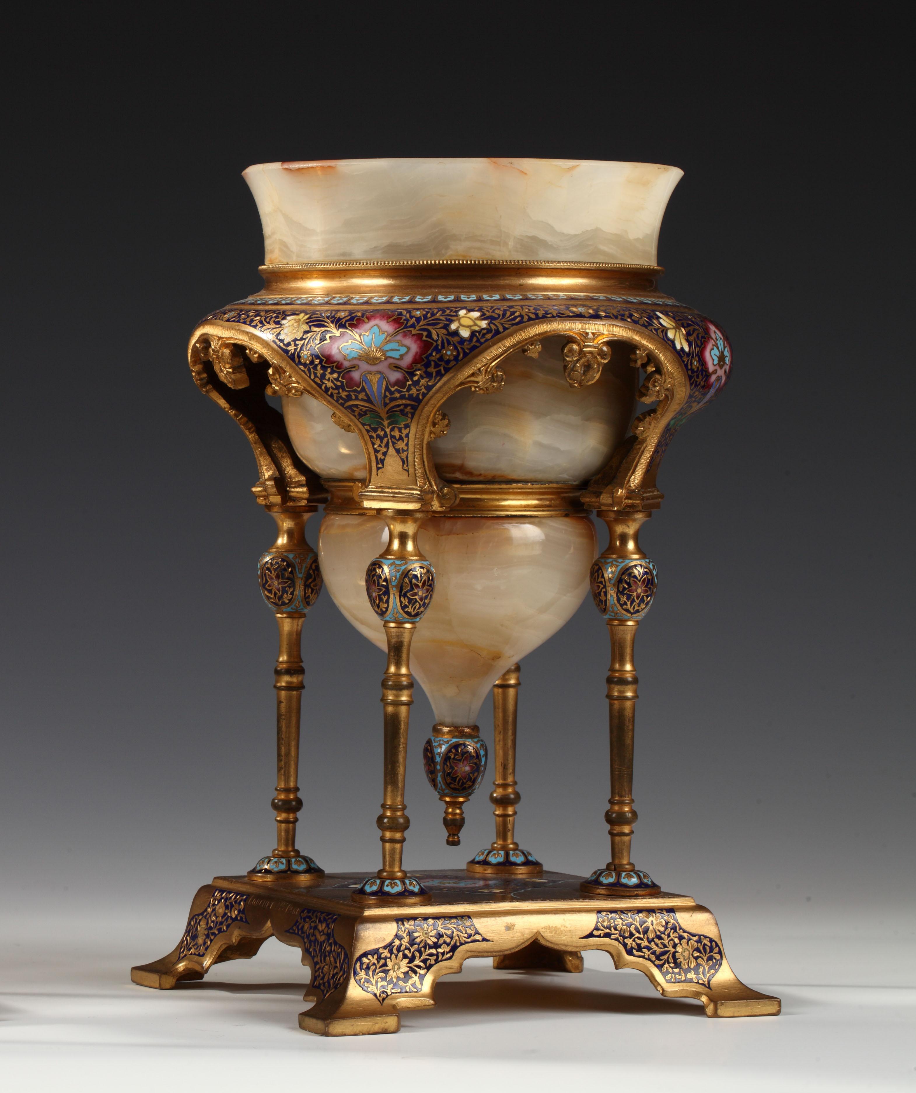 Élégante coupe orientale en onyx attribuée à Louchet Frères, insérée dans une monture architecturale en bronze doré sur quatre colonnes supportant une large moulure enrichie de motifs feuillagés en émail cloisonné polychrome. L'ensemble repose sur