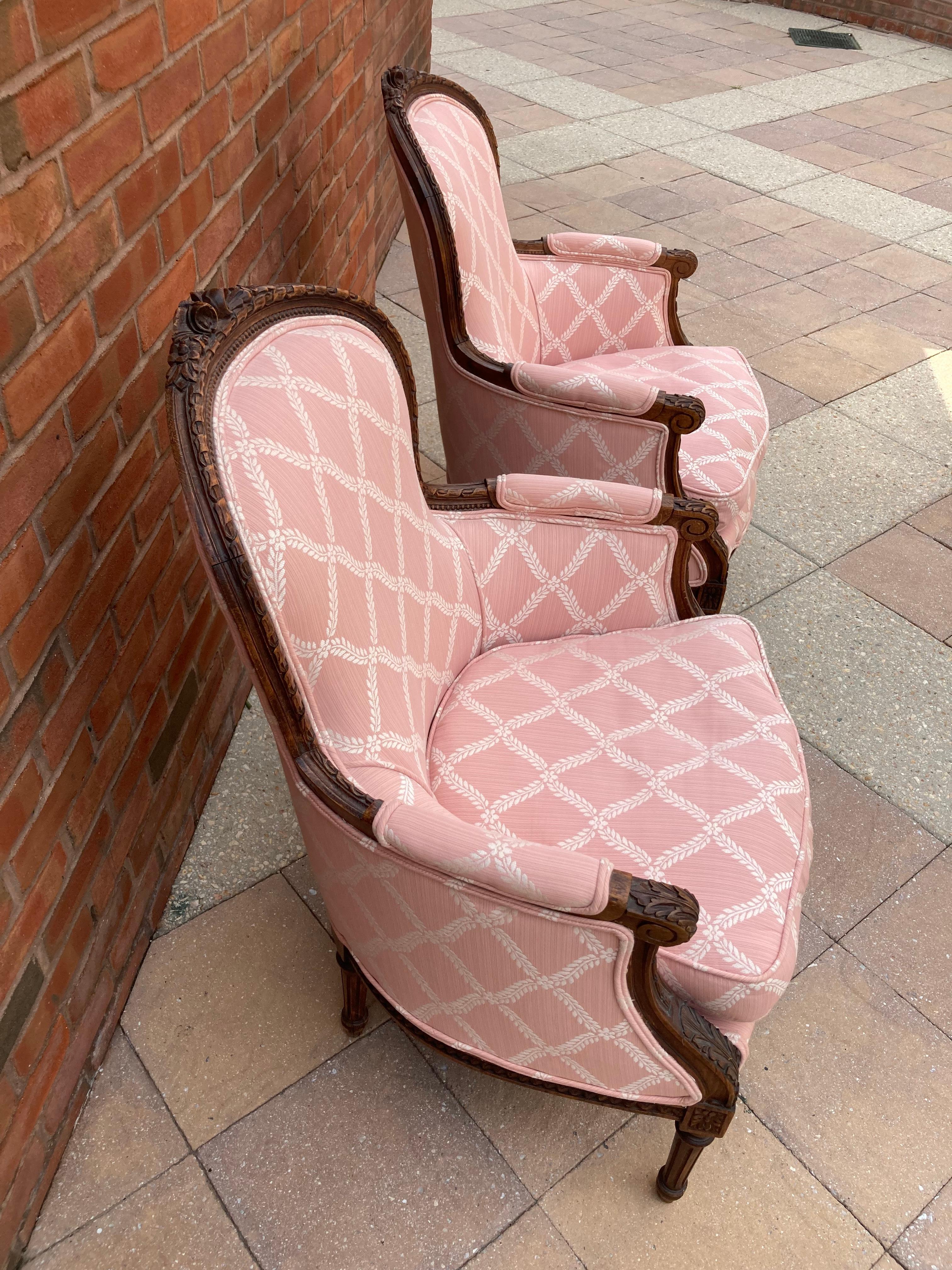 Beautiful upholstered pair of French styled bergere chairs with fully carved wood frames featuring acanthus leaves, fluted legs and flowers. White on pink fabric is in a diamond pattern. Seat slip covers are removable. Cushions are down and feather
