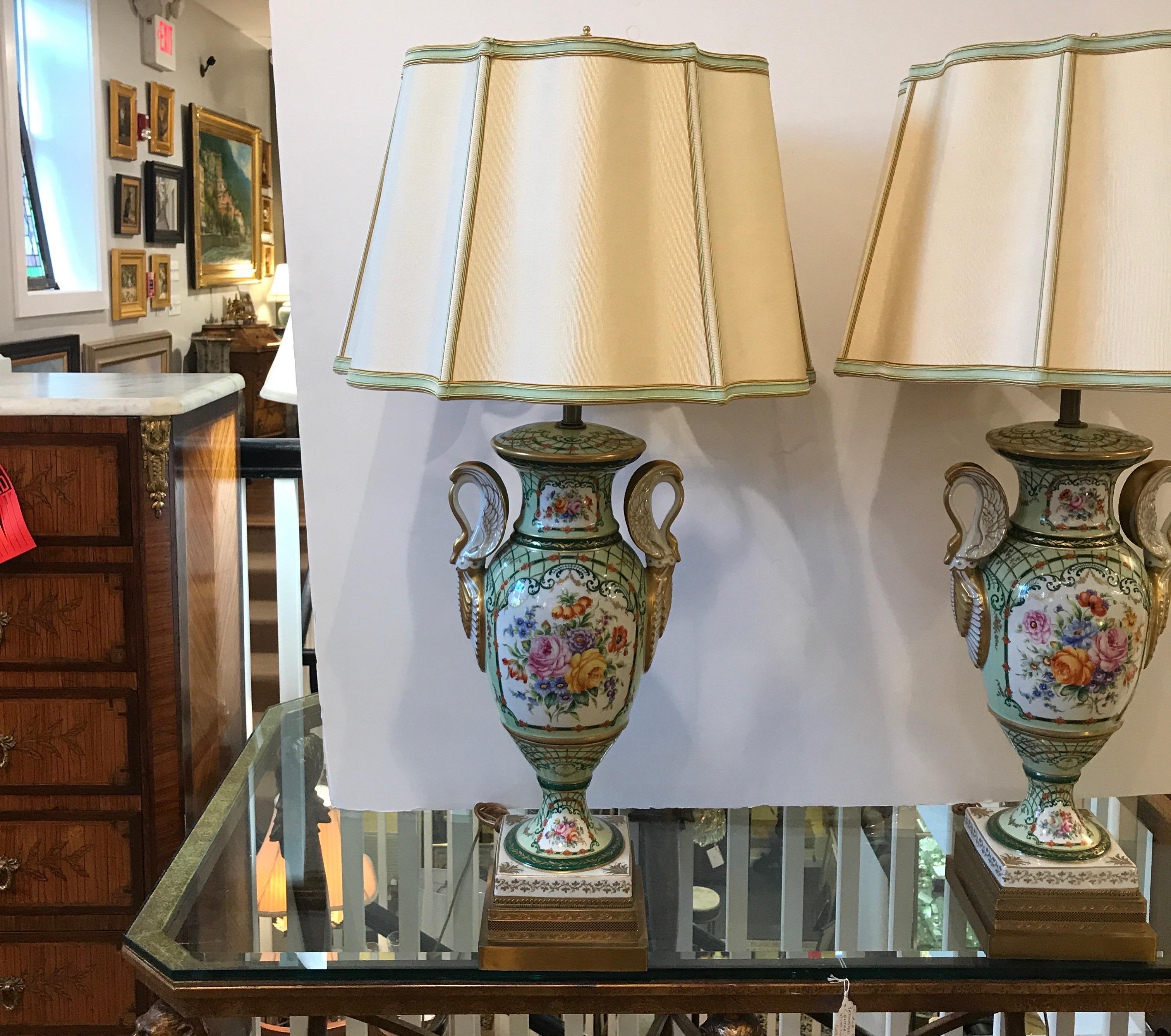 A pair of French porcelain hand painted urn form lamps with custom silk taffeta shades. The highly decorated lamps with swan gilt handles and bouquets of flowers on both sides of the urns and bases. The lamps each have double sockets with pull chain