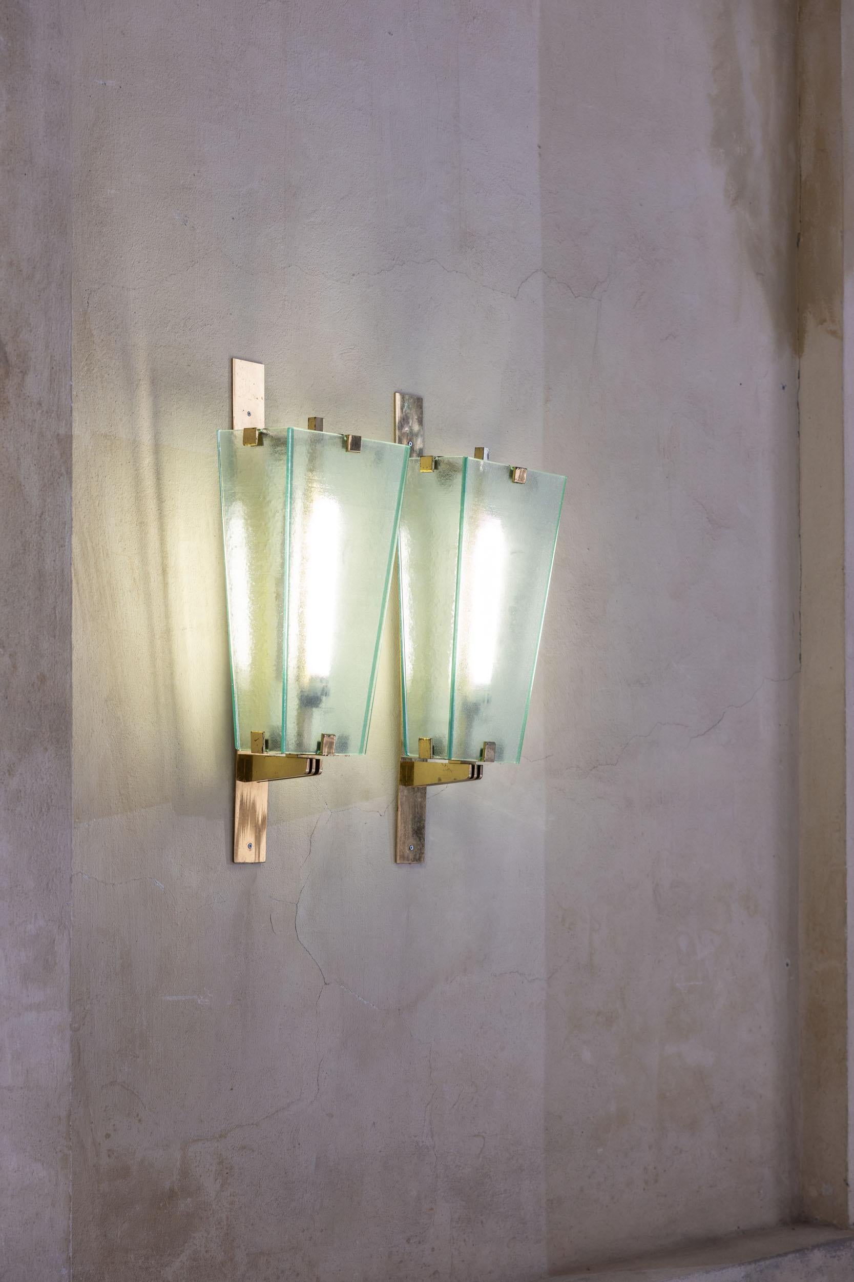Elegante coppia di lampade da parete, caratterizzate da un morbido vetro sfumato color verde acqua e da una struttura geometrica in ottone che ne esalta l'eleganza senza tempo. La combinazione di vetro delicato e ottone robusto crea un contrasto