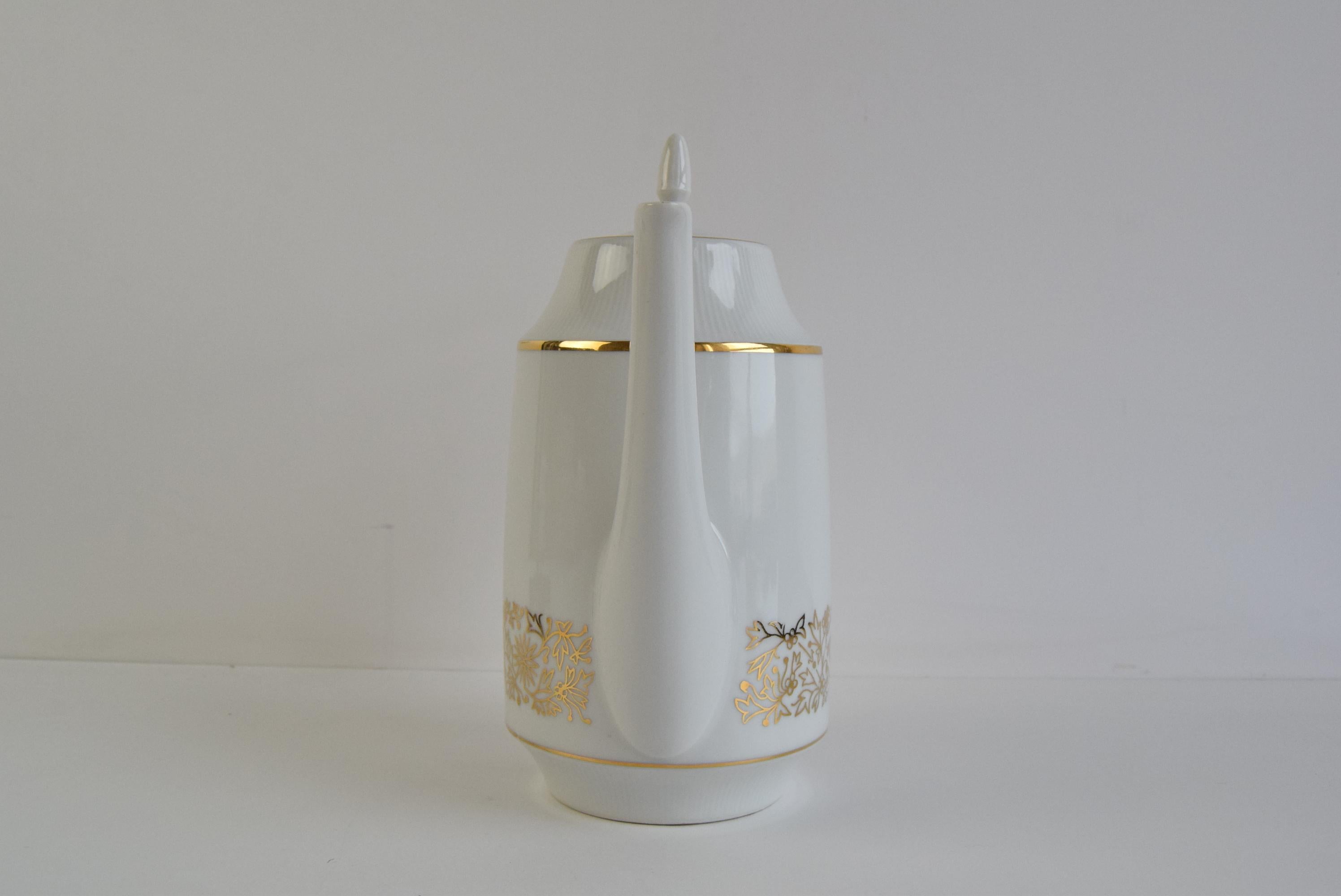This mid-century piece features a sleek white body adorned with refined gold floral motifs and a gold accent band around the top. Its sharp, modernist handle and spout give it a sophisticated yet functional form typical of late 1960s European