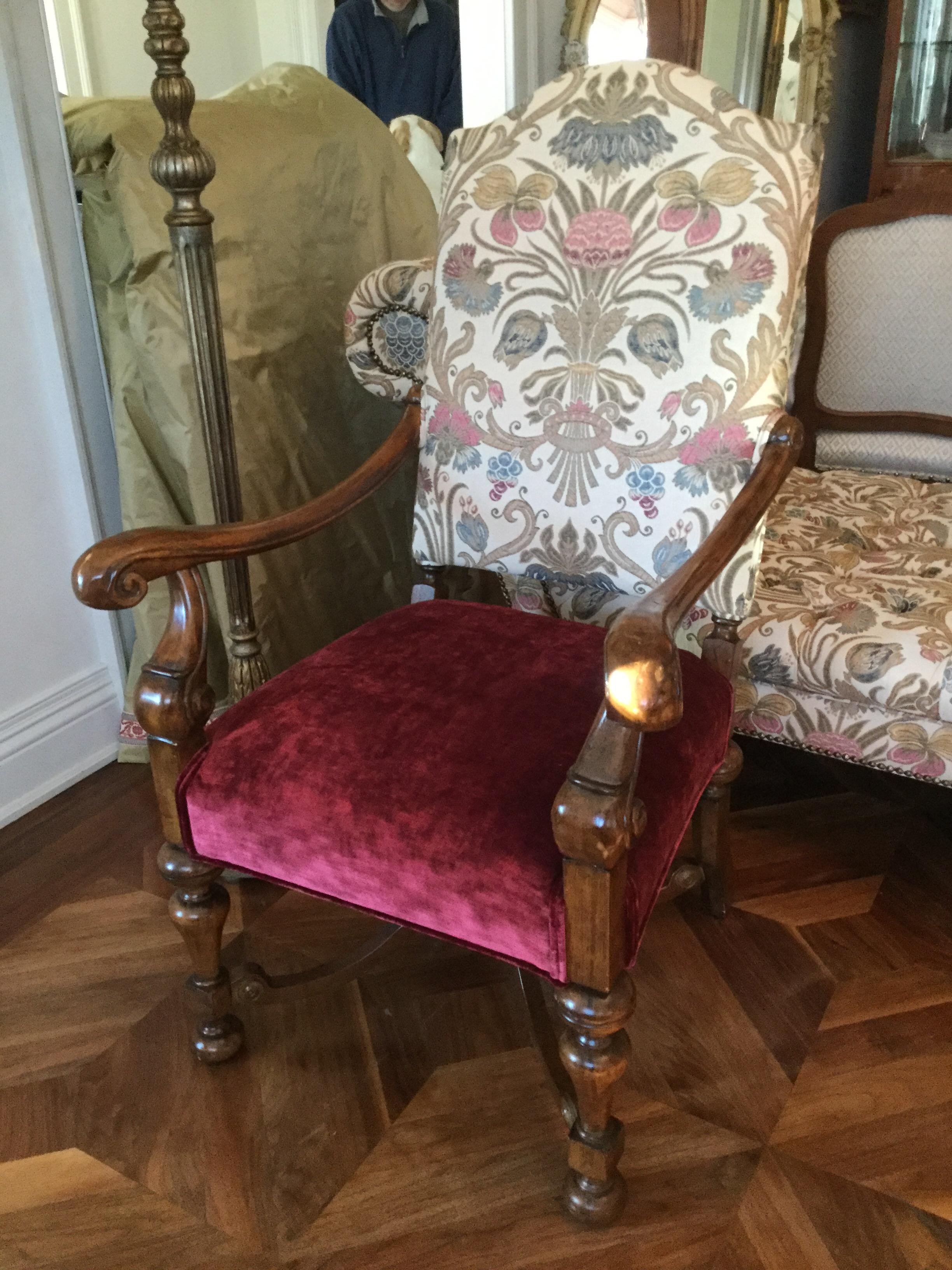 Elegantly carved wood frame featuring Renaissance-style legs, elongated sweeping arms, and a cross-stretcher for stability. This high-back chair is upholstered in a rich burgundy velvet seat and a patterned back in shades of pink, blue and beige.