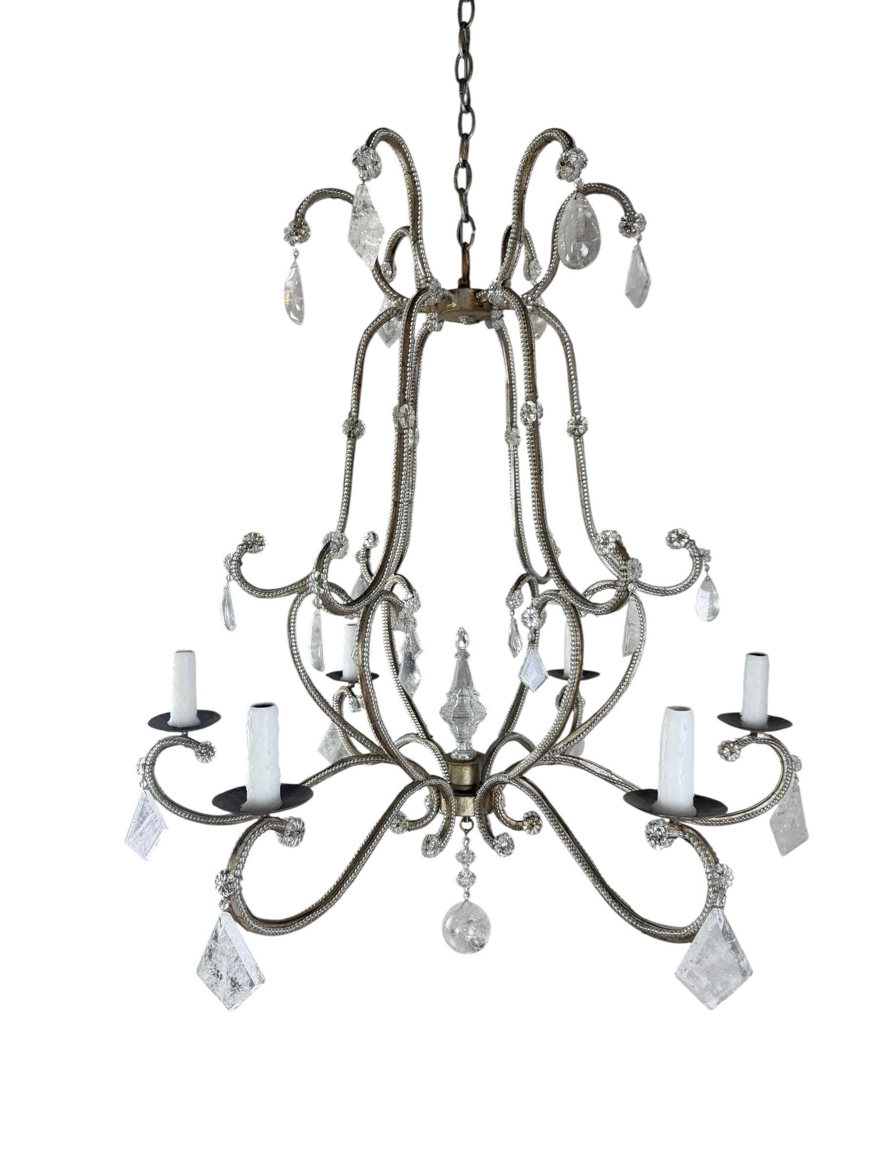 Un élégant lustre à huit lumières composé de bras en fer à volutes enveloppés de perles de cristal, ornés de pendentifs en cristal de roche et en cristal facetté. Le lustre présente un cadre aux contours gracieux avec des volutes, culminant avec un