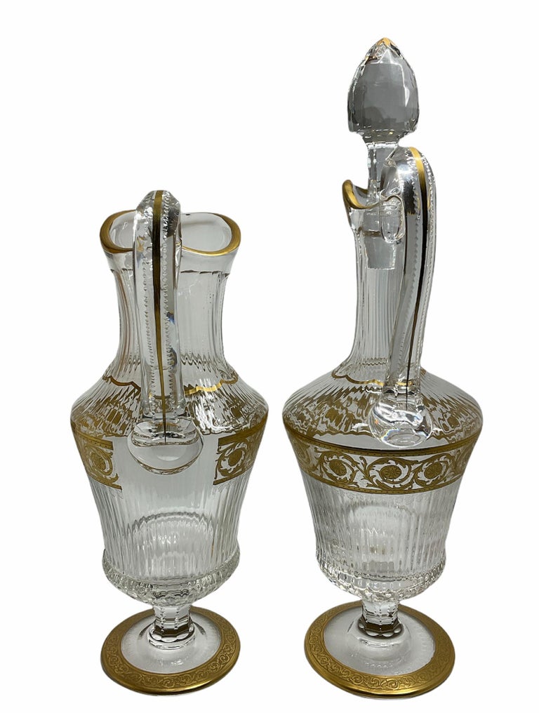This is a set of Saint Louis crystal gold Thistle pattern of a jug and decanter with handles. Their crystal are adorned with vertical fluting and cutting decoration leaves. Also, a band of scrolls of gold thistle branches decorate their upper body