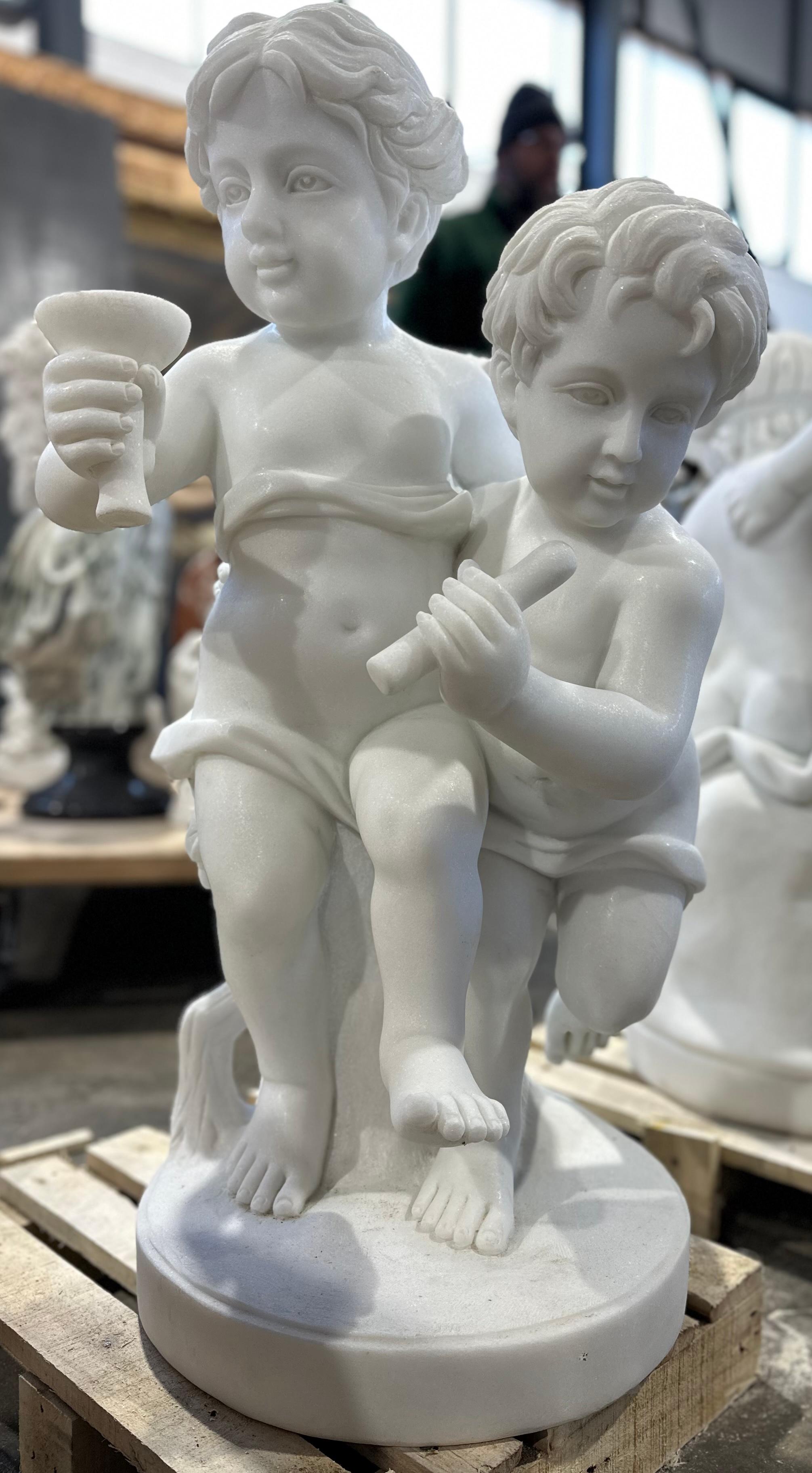 Eine elegante Skulptur aus weißem Marmor mit zwei kindlichen Putten, die fröhlich spazieren gehen, während die eine aus einem Becher trinkt und die andere sich an einen Ast lehnt. 
Die Schnitzerei ist klar und detailliert, von den gewellten Haaren