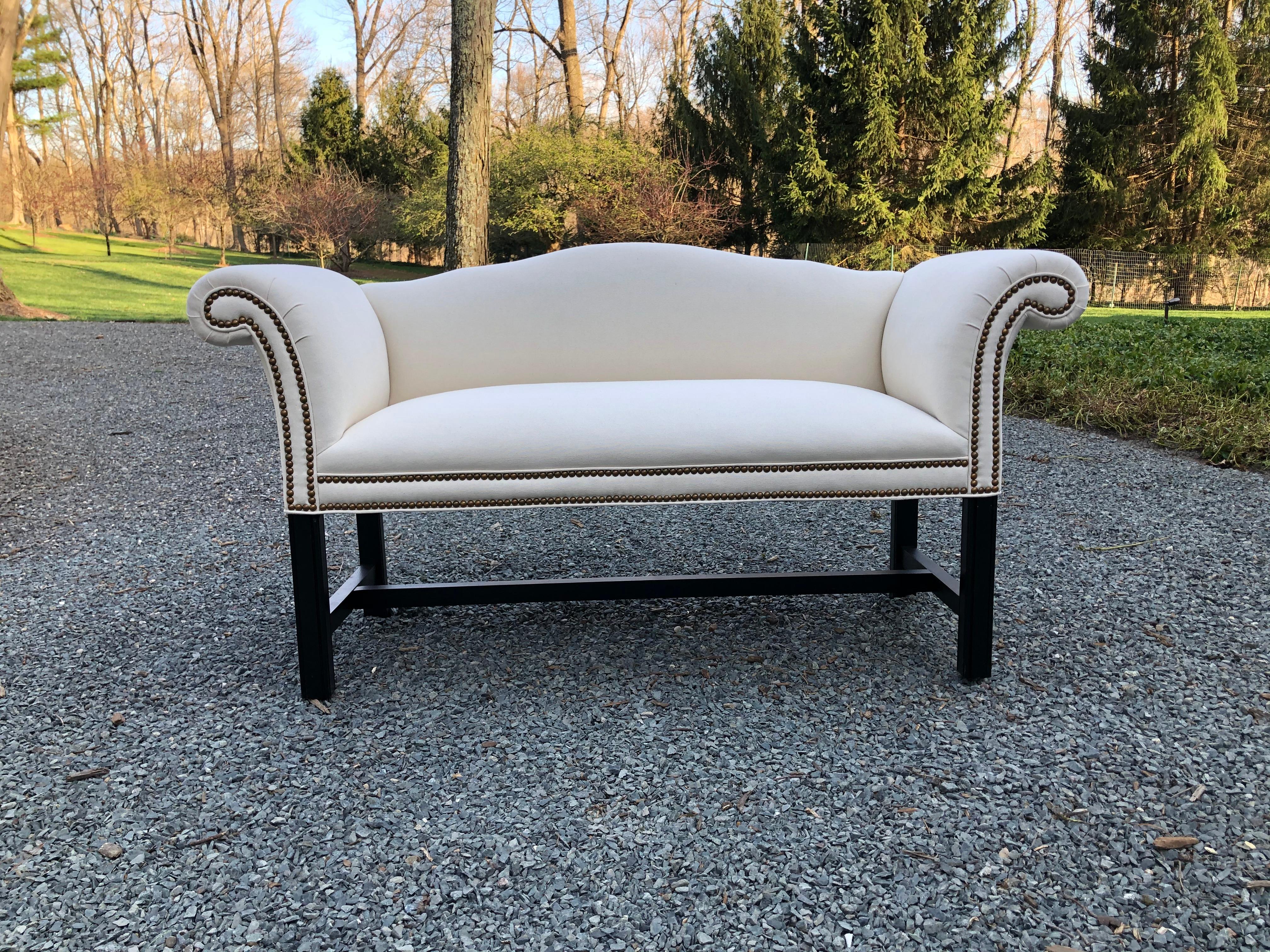 A vintage camelback settee newly upholstered in a crisp white cotton duck and adorned with antiqued French nailheads having scrolled arms and ebonized legs and cross stretcher.