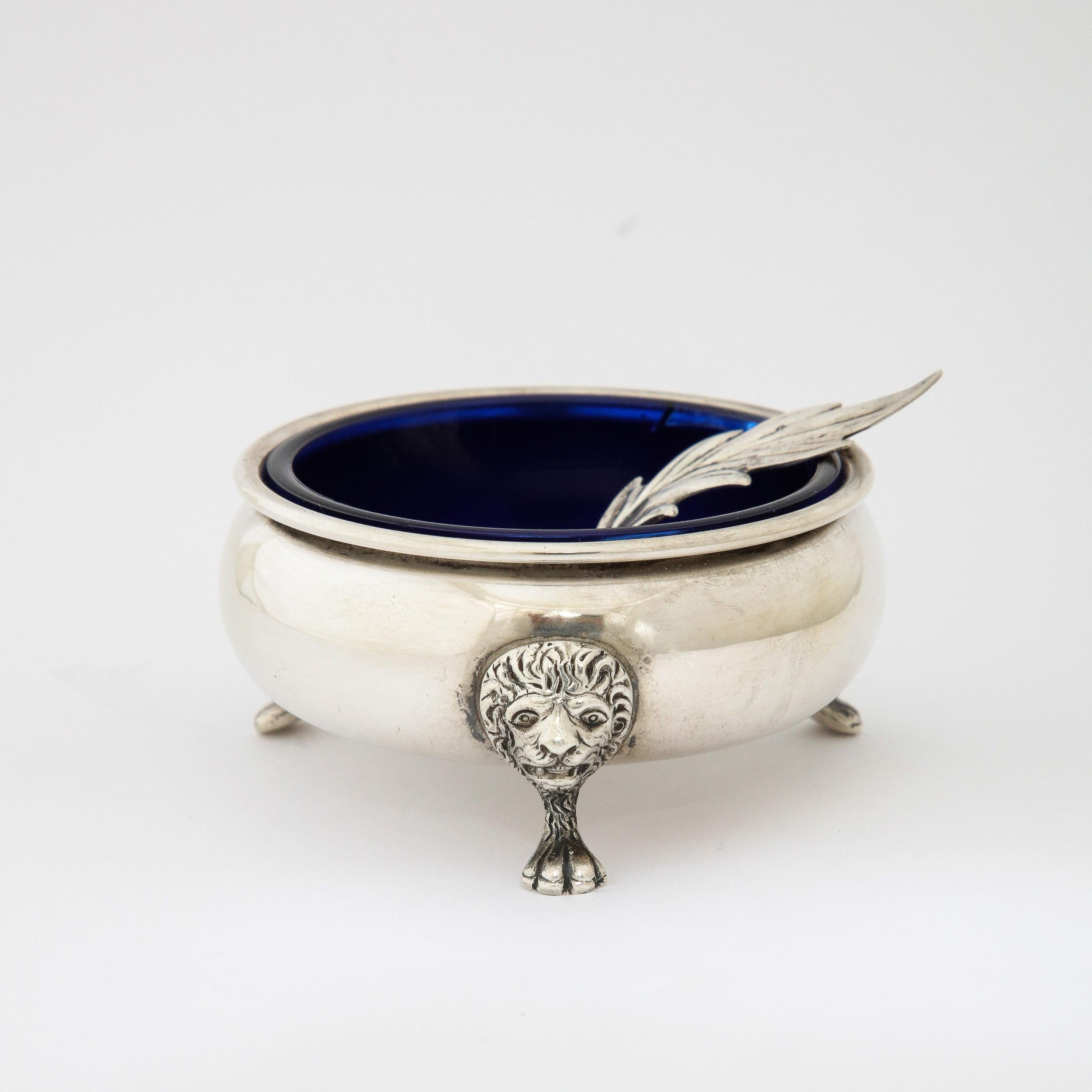 This very elegant Neoclassical sterling silver salt cellar features three stylized lion and paw feet and this piece is also inset with an original cobalt glass liner. It also has a stylized leaf spoon as well that is also marked sterling. Very good