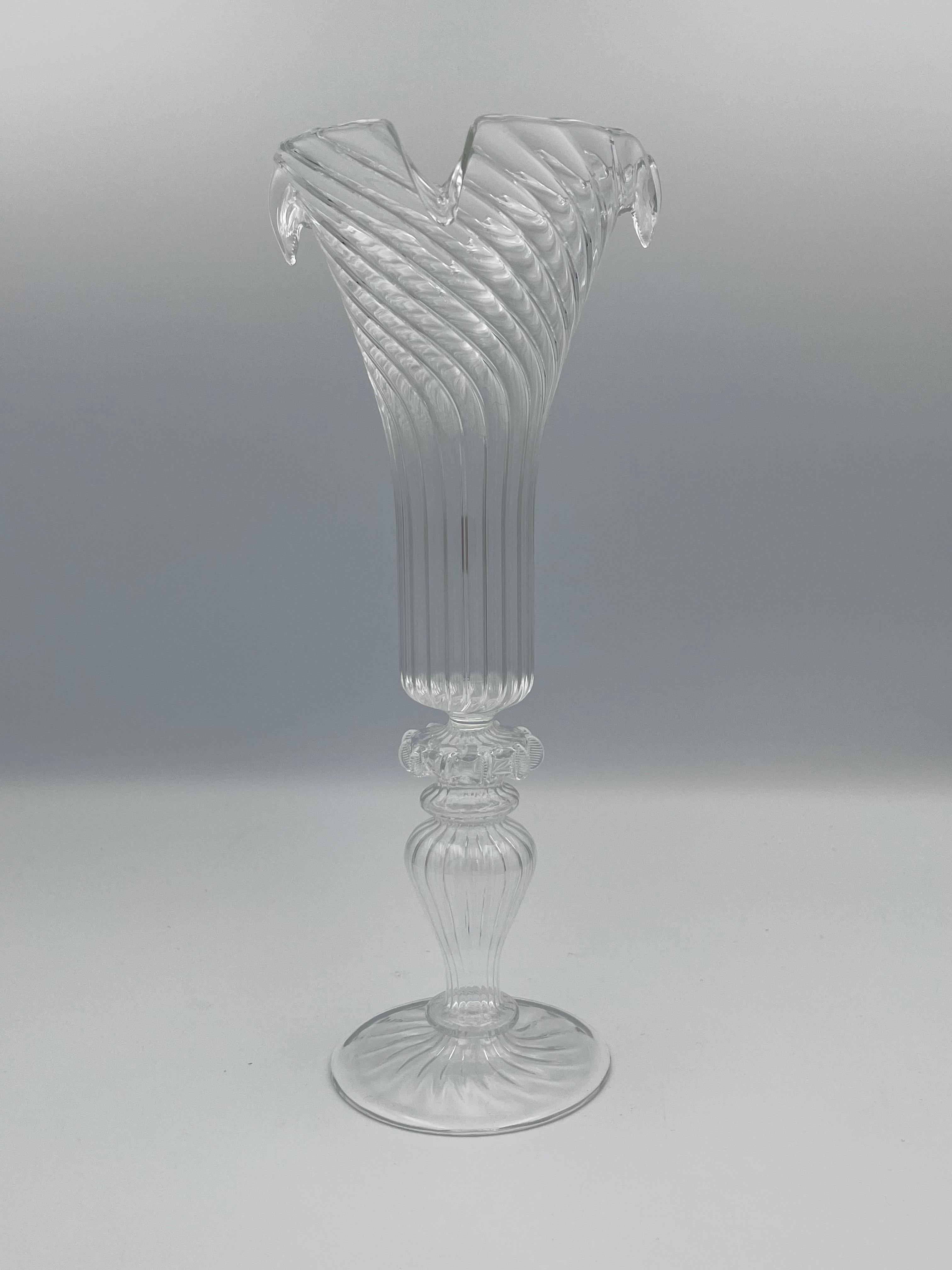 Este exquisito jarrón de cristal transparente soplado a mano se alza sobre una escultural base de pedestal, mostrando una artesanía magistral y una forma elegante y fluida. El cuerpo presenta un distintivo diseño acanalado que se retuerce hacia